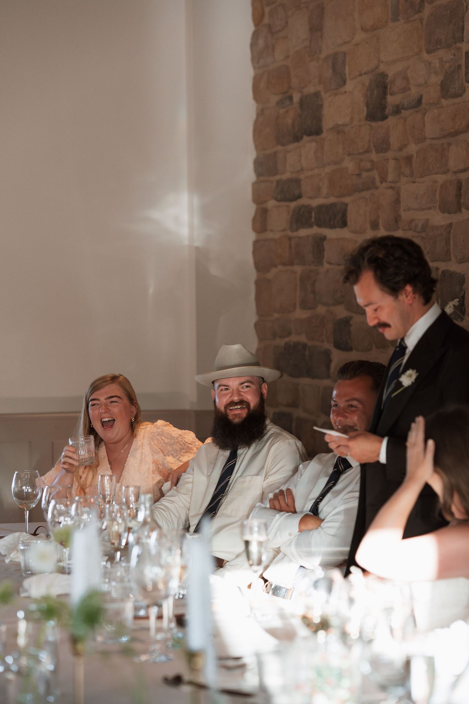 Wedding party members laughing together at reception head table against brick wall.