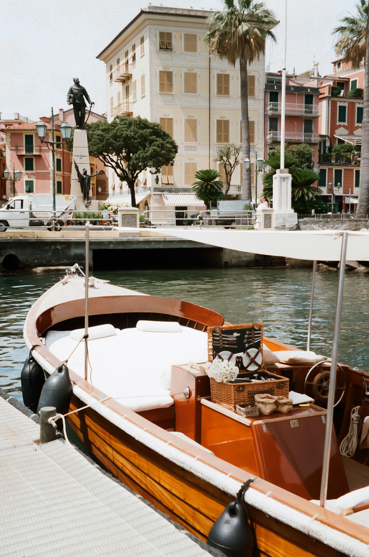 Wooden boat with polished mahogany hull docked at Mediterranean waterfront near white buildings.