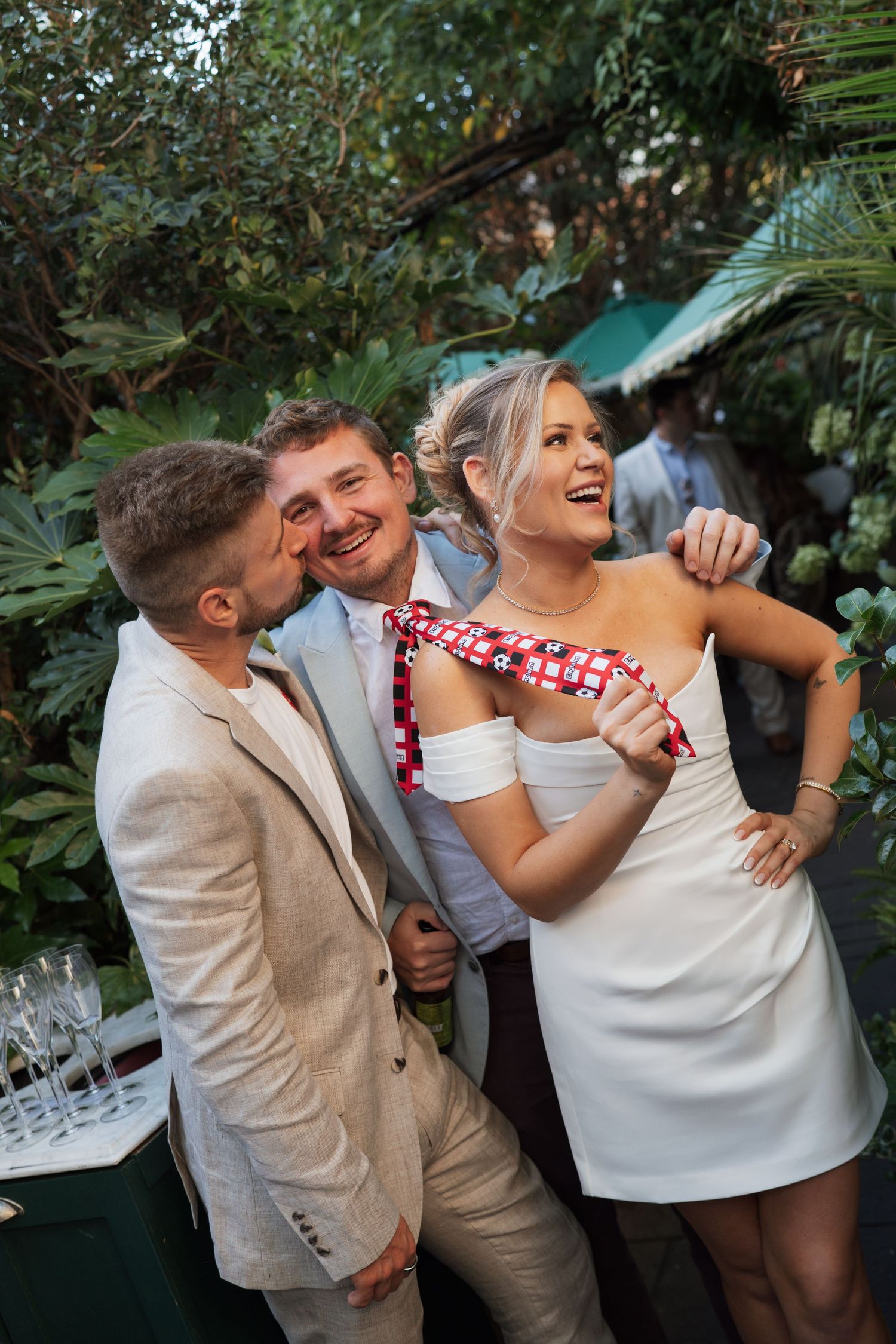 Two men and a woman in white dress laugh together at an outdoor wedding reception surrounded by lush greenery.