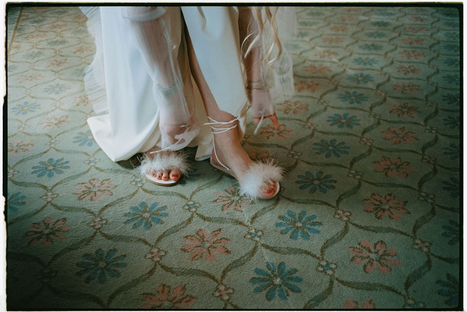 Bride wearing feathered white heeled sandals on a vintage floral patterned carpet.