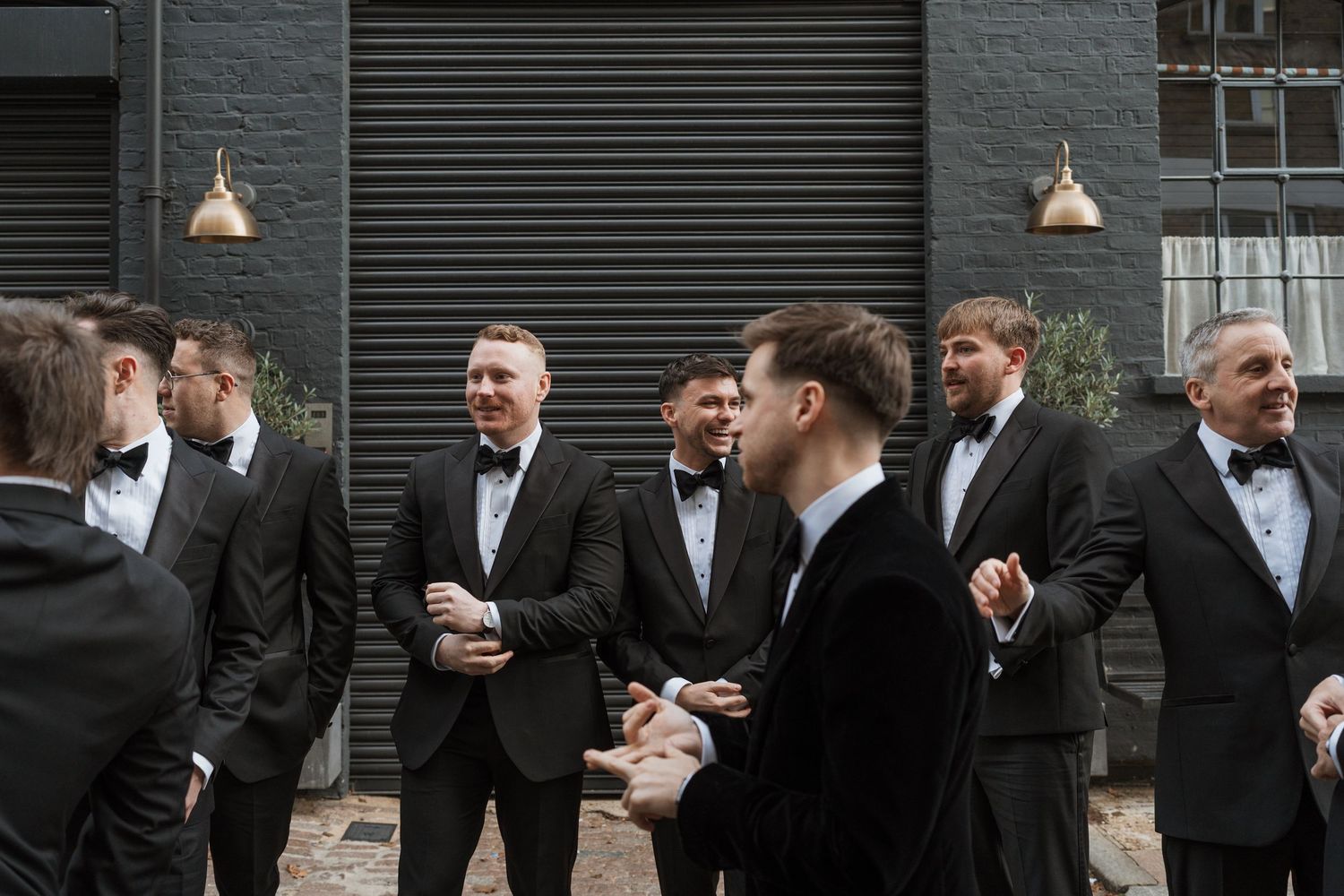 Group of groomsmen in black tuxedos gathered outside for casual conversation.