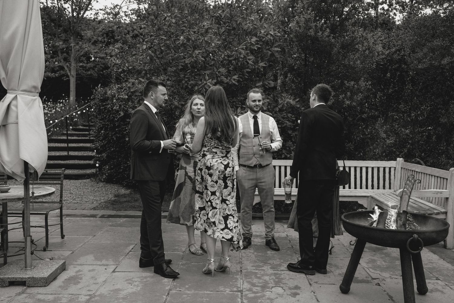 Outdoor gathering with people mingling on garden path near white benches.