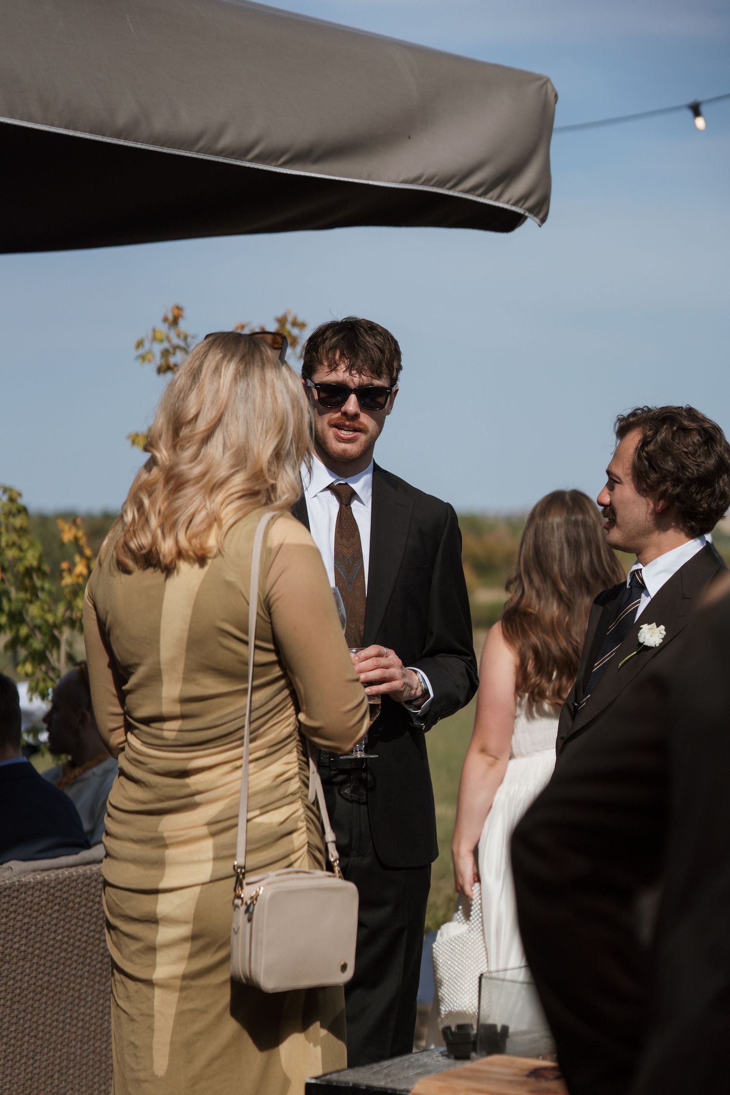 Guests mingling at an outdoor reception with drinks against a sunny backdrop.