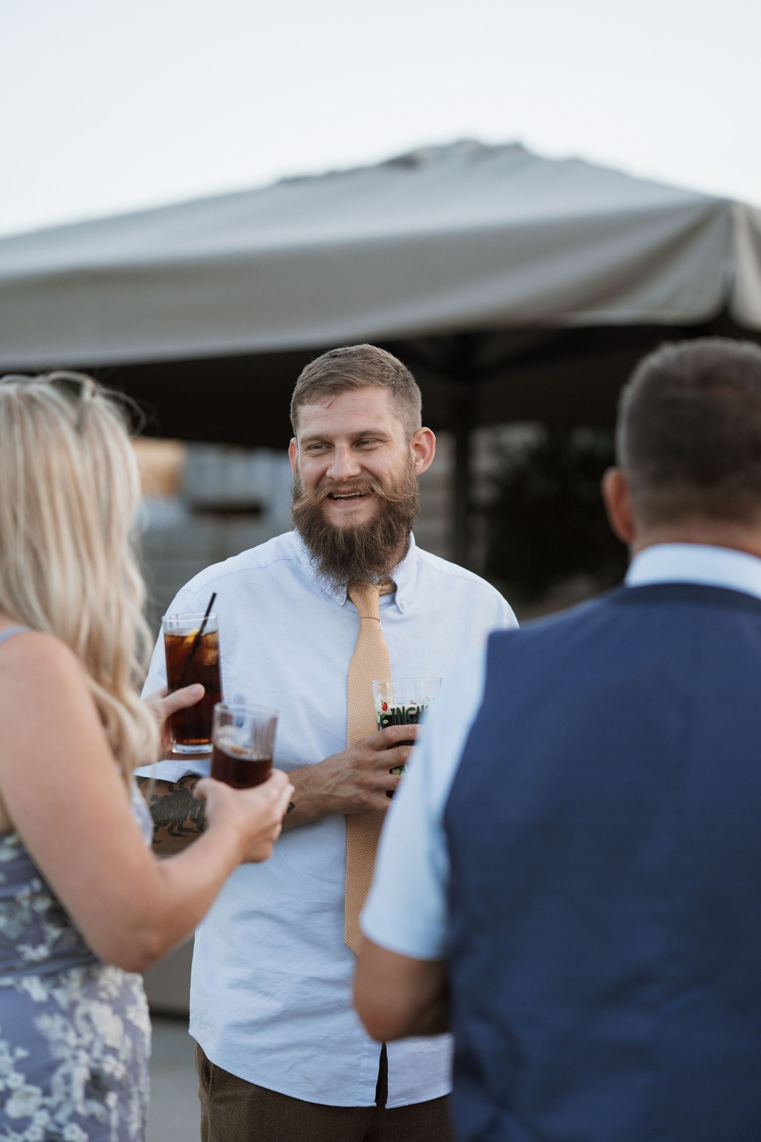 Guests mingling and holding drinks under a white tent at an outdoor event.