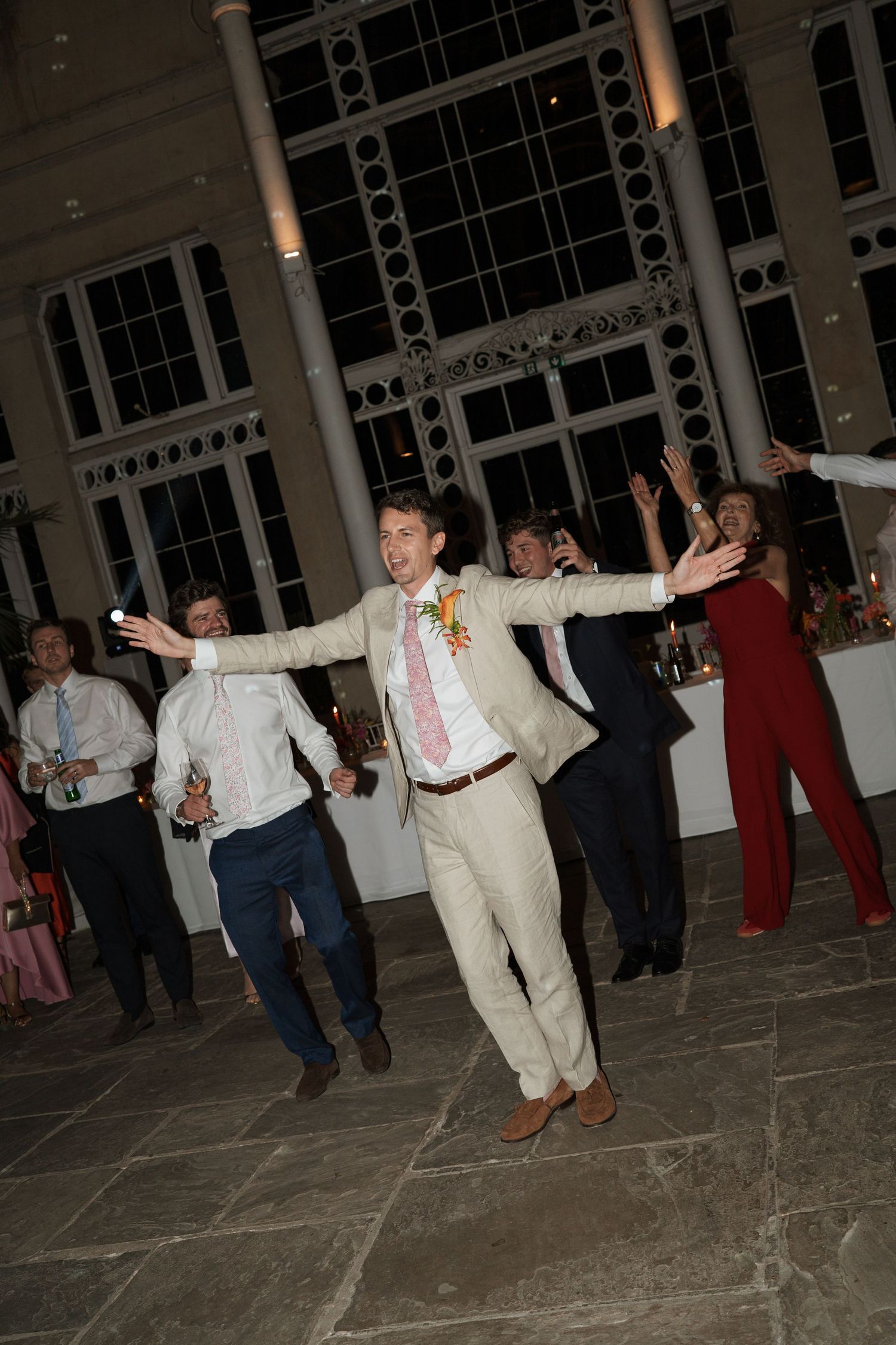 Wedding guests dancing in a line on a tiled floor during reception celebrations.