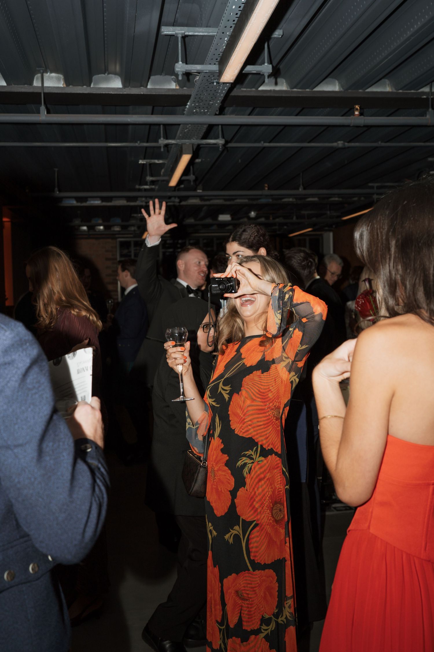 Party guests in colorful dresses enjoying drinks and dancing under industrial lighting.