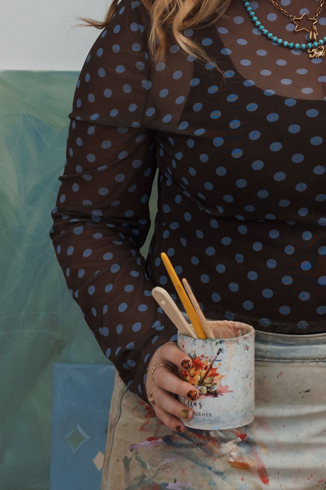 Artist holding paint brushes in a cup, wearing a brown polka dot mesh top and paint-splattered apron.