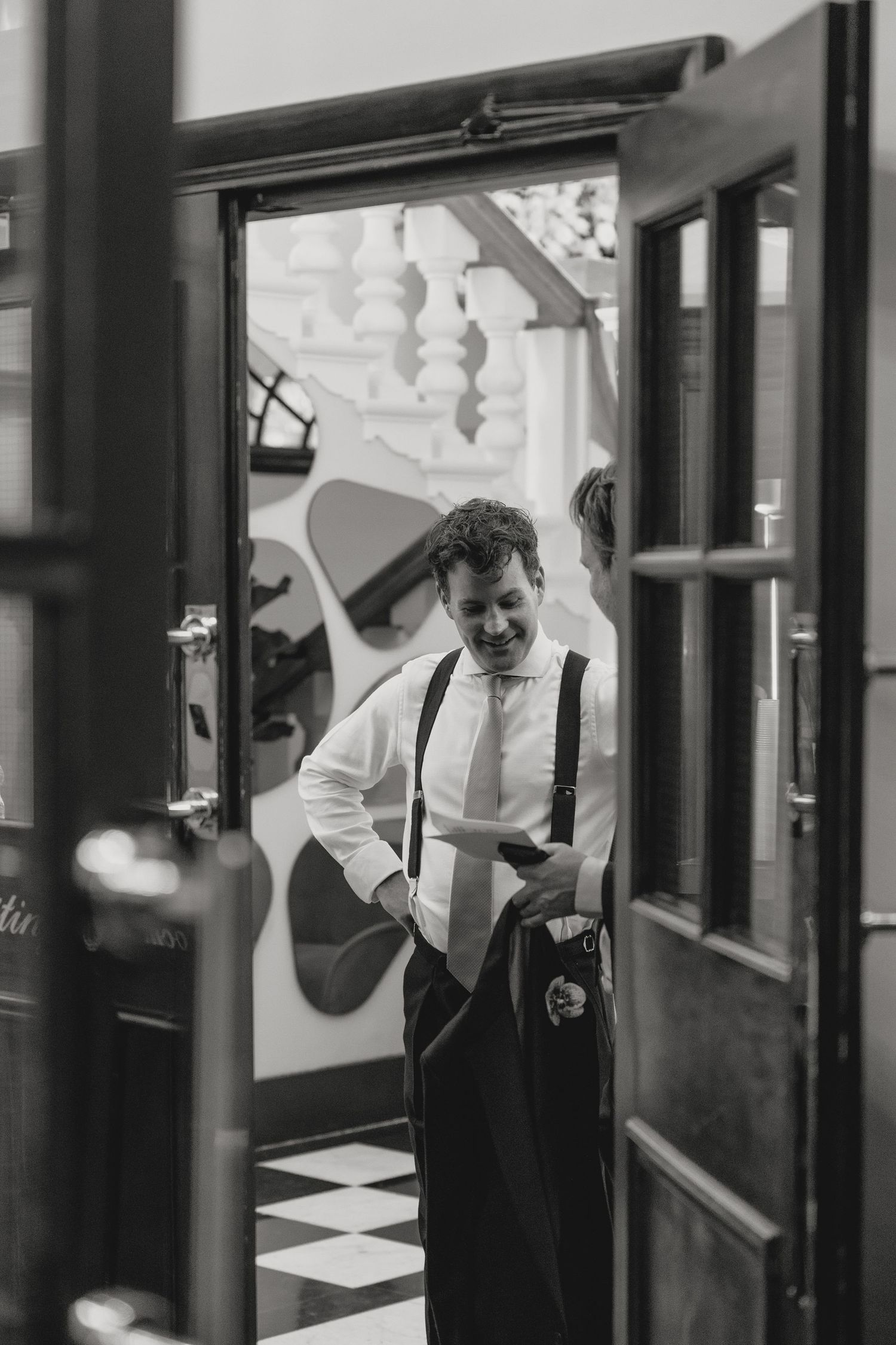 Black and white photo of someone in formal attire with suspenders seen through a doorway.