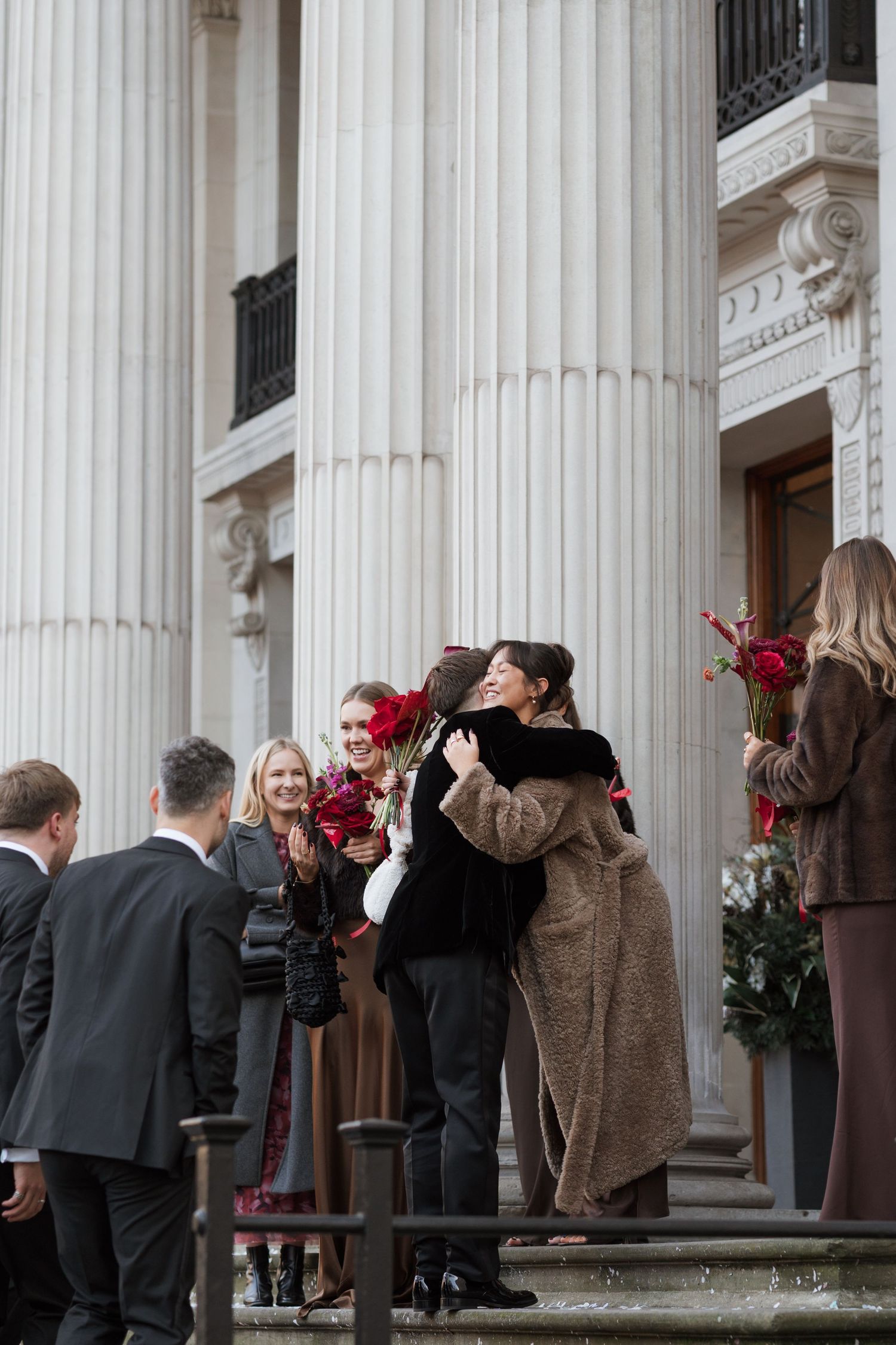 A crowd gathers outside a building with white classical columns and red flowers.