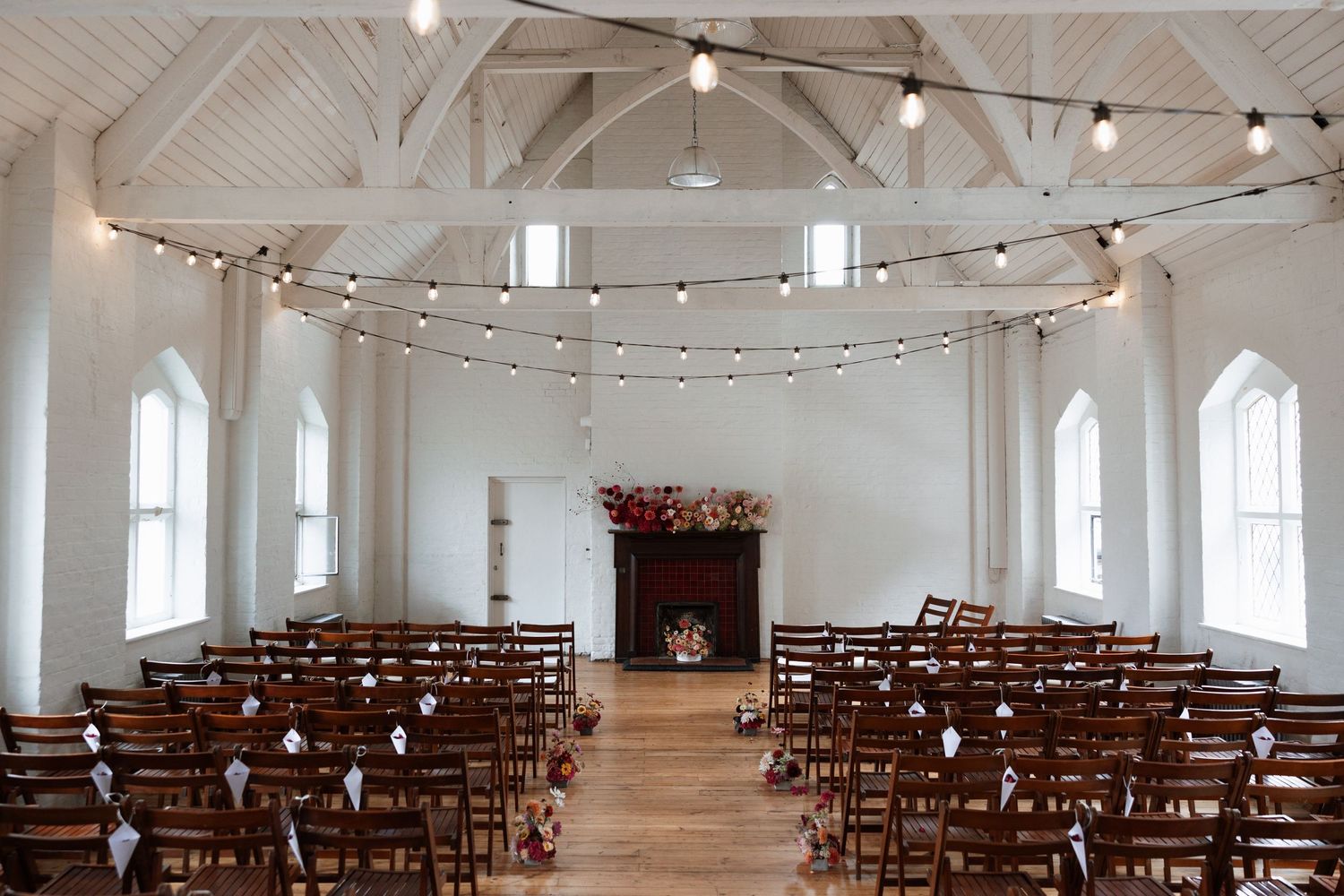 Rustic wedding ceremony venue with wooden chairs in two aisles, string lights overhead, a fireplace with floral display, and arched windows.