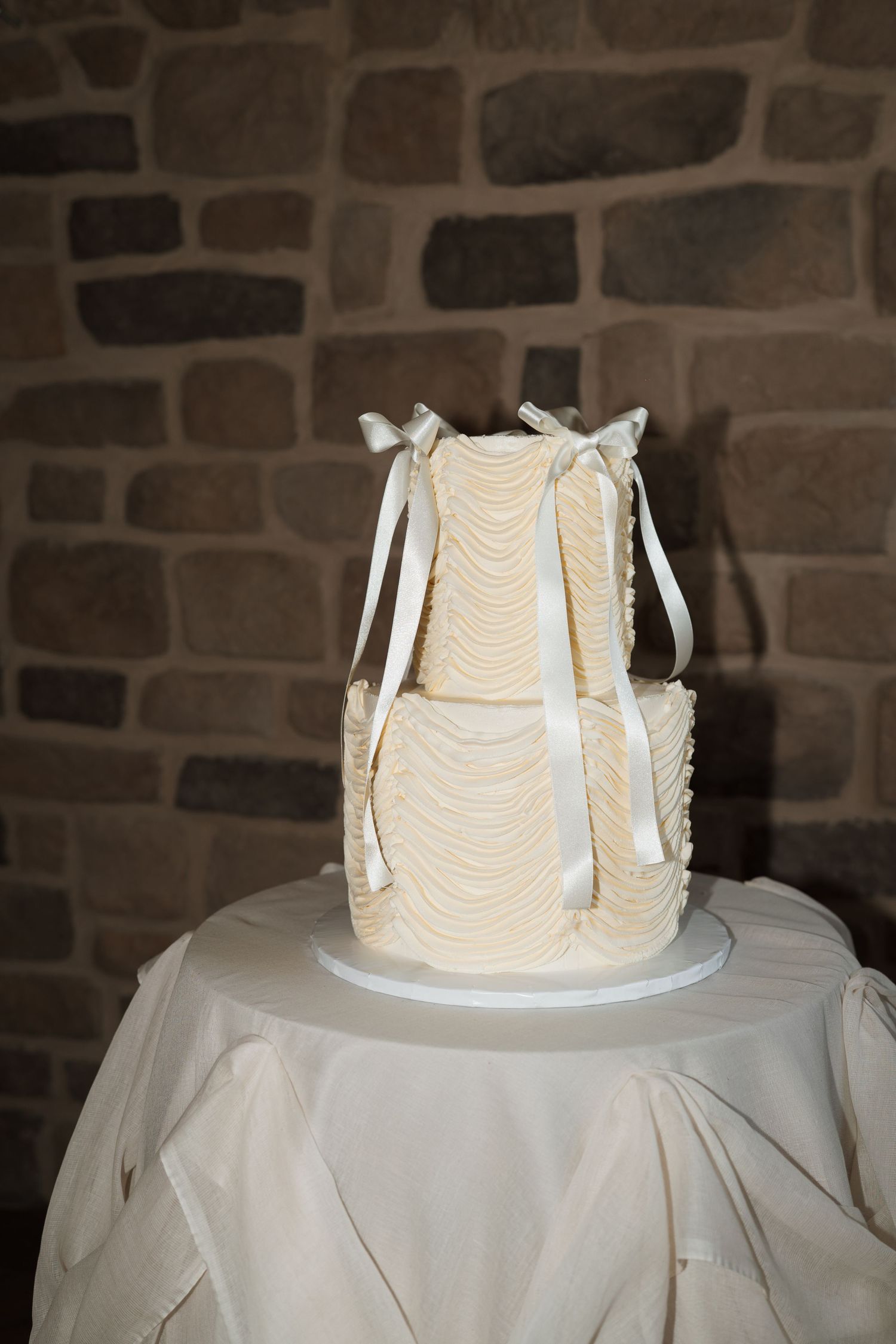 Close-up of a white wedding cake with ribbon details against a rustic stone wall.