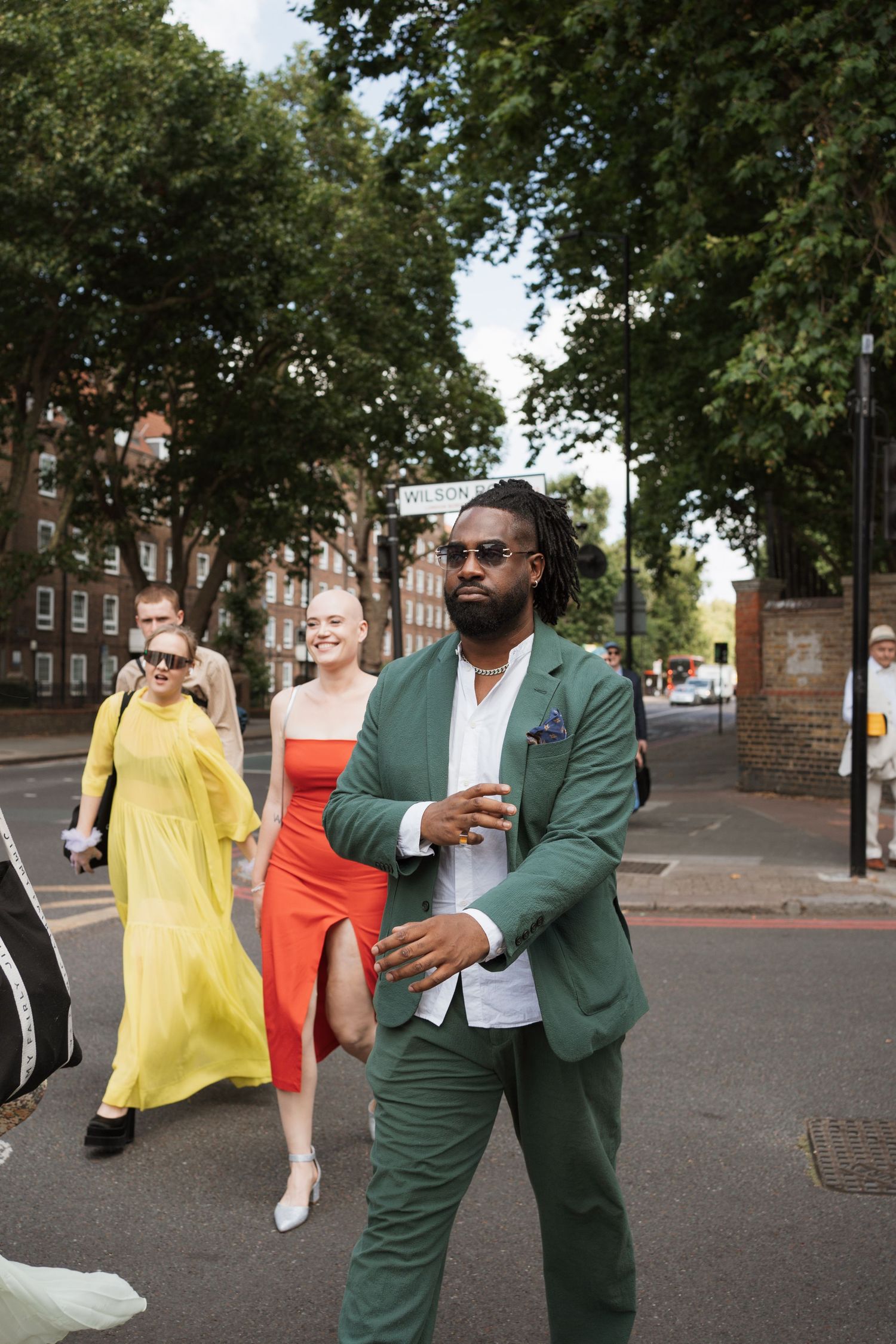 Wedding party members in bright dresses and suits walking together on a tree-lined street.