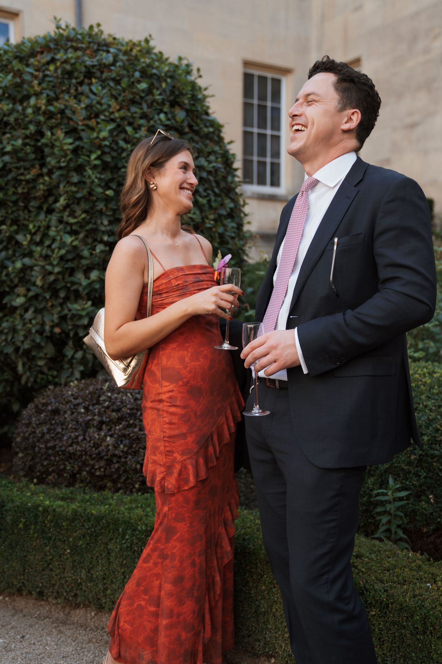 Couple shares a candid moment of laughter while holding drinks at an outdoor wedding reception.