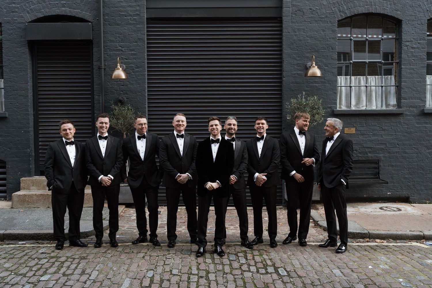 Wedding party in formal attire poses against dark brick wall outdoors.