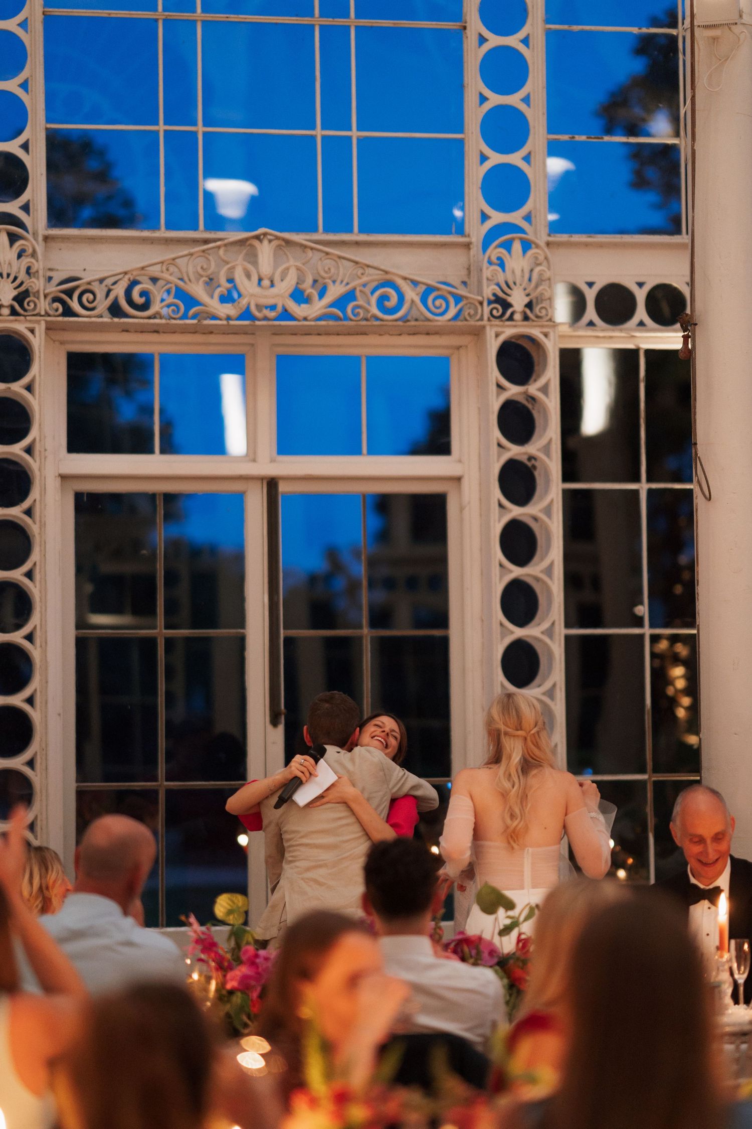 Large ornate windows frame an elegant reception dinner under blue evening sky.