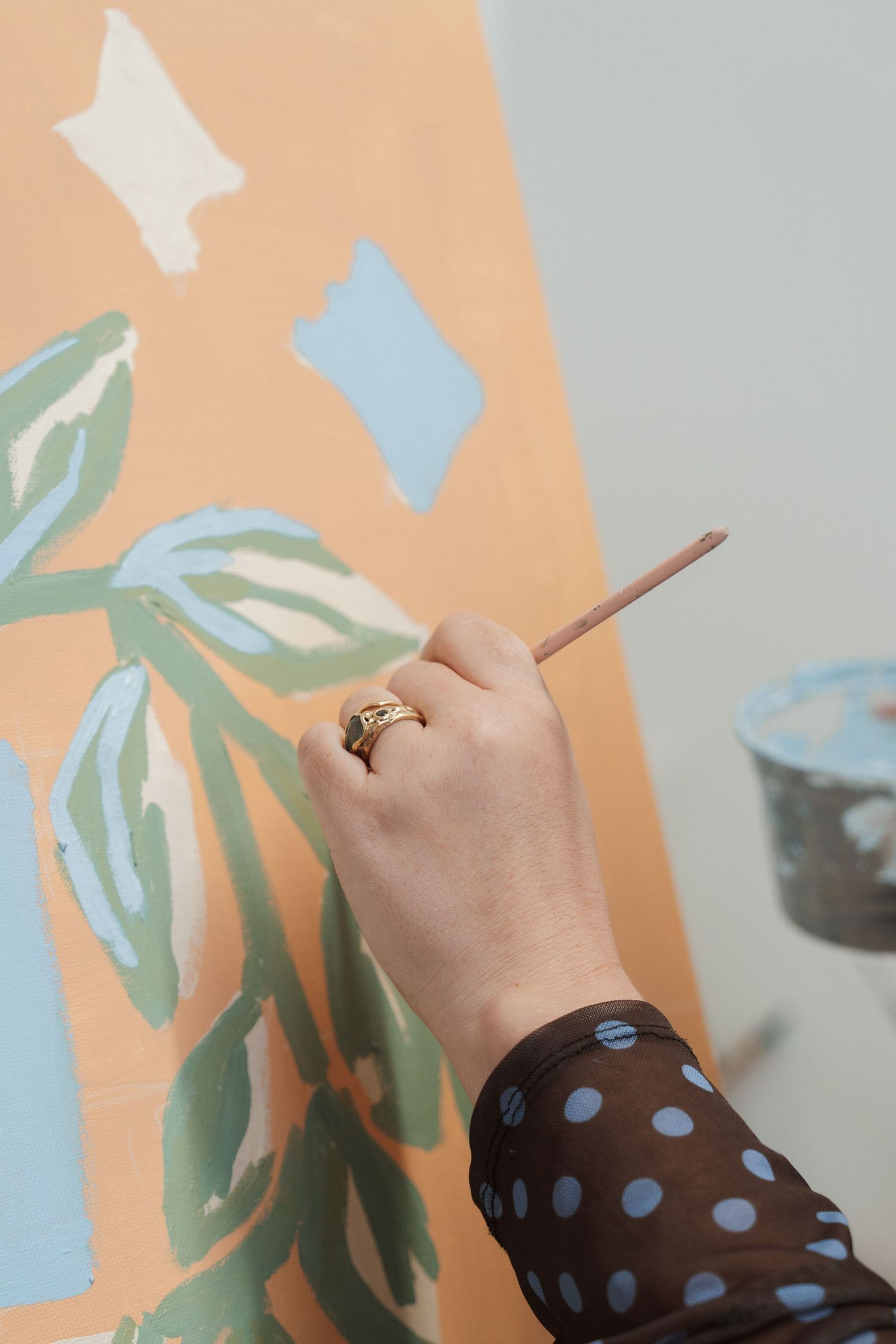 A hand wearing a gold ring painting a colorful mural with green leaves and blue shapes on an orange wall.