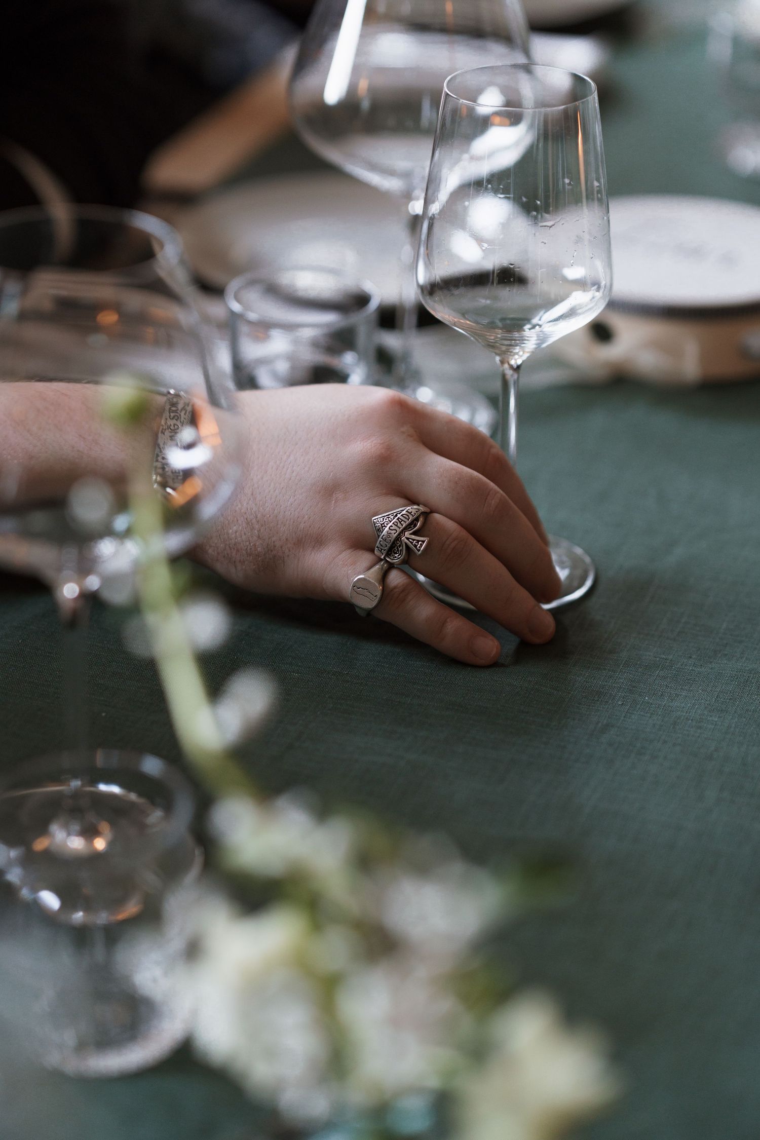Close-up of hands near wine glasses on dark green tablecloth with white flowers.
