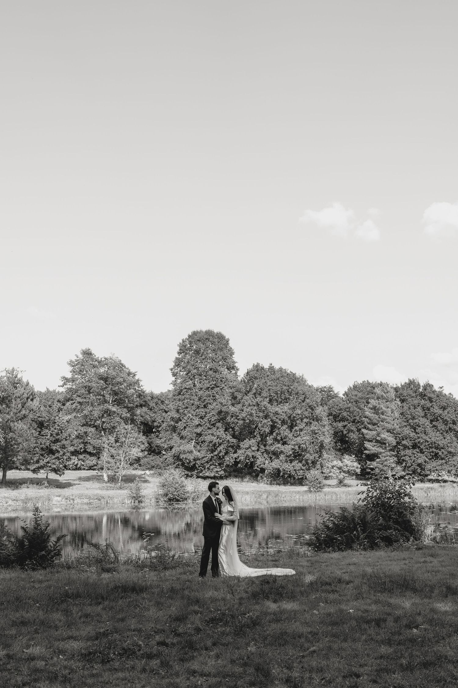 A black and white wedding portrait with a long flowing veil against a pastoral landscape.
