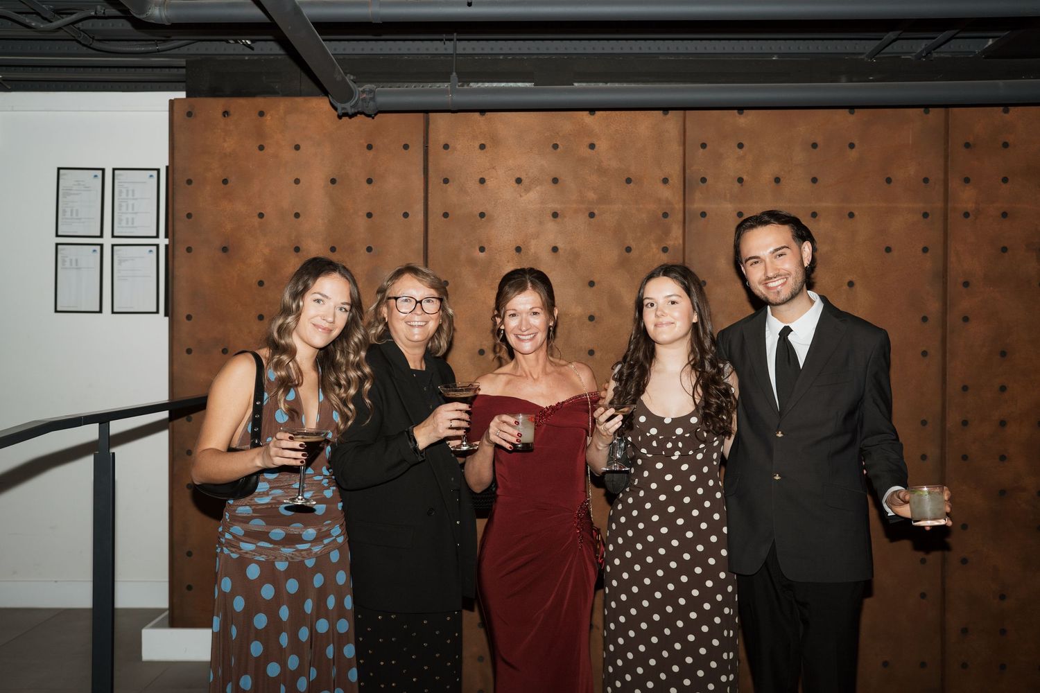 Group of party guests posing together against a wooden paneled wall at an evening event.