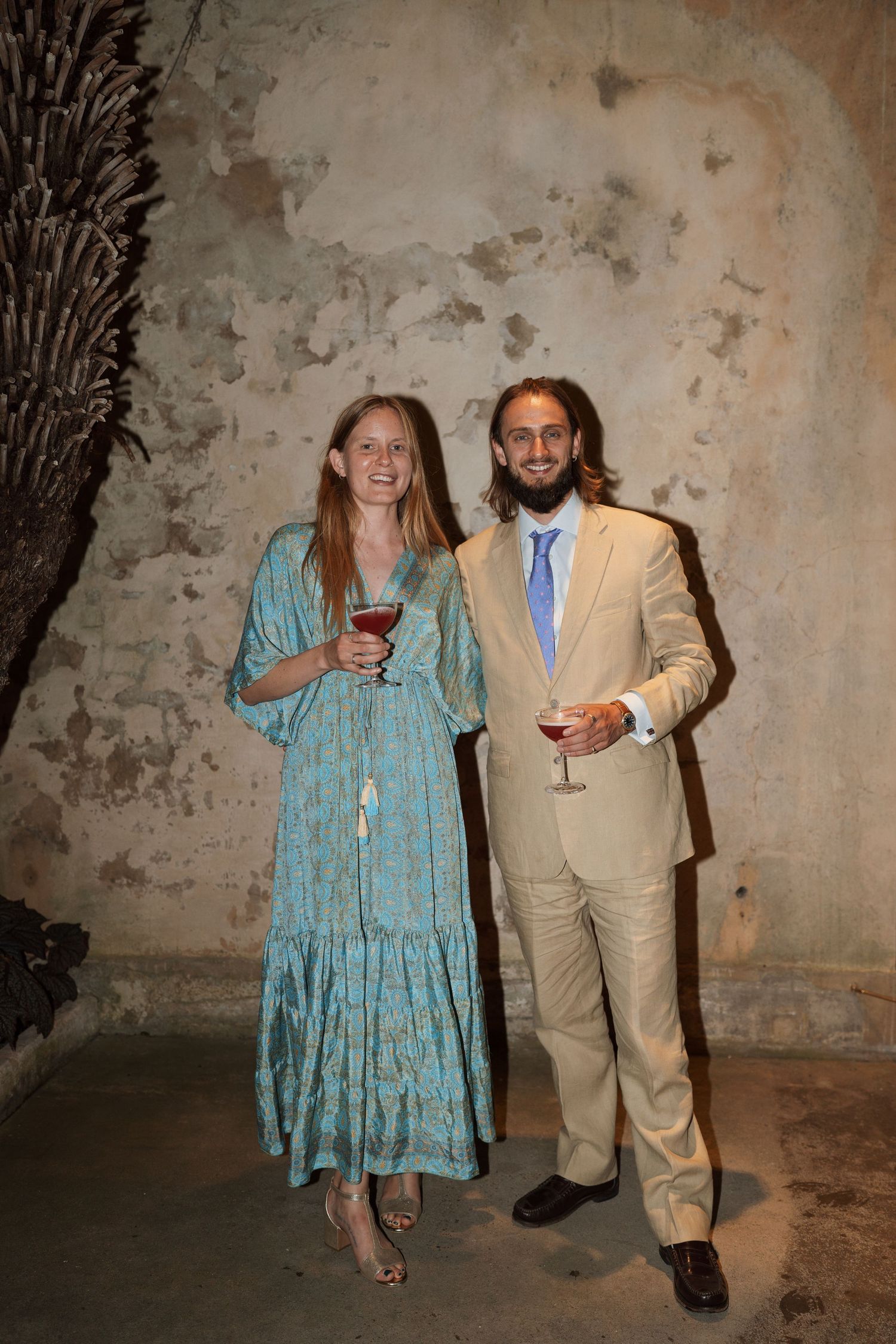 Wedding guests in formal attire pose against a weathered wall with drinks.
