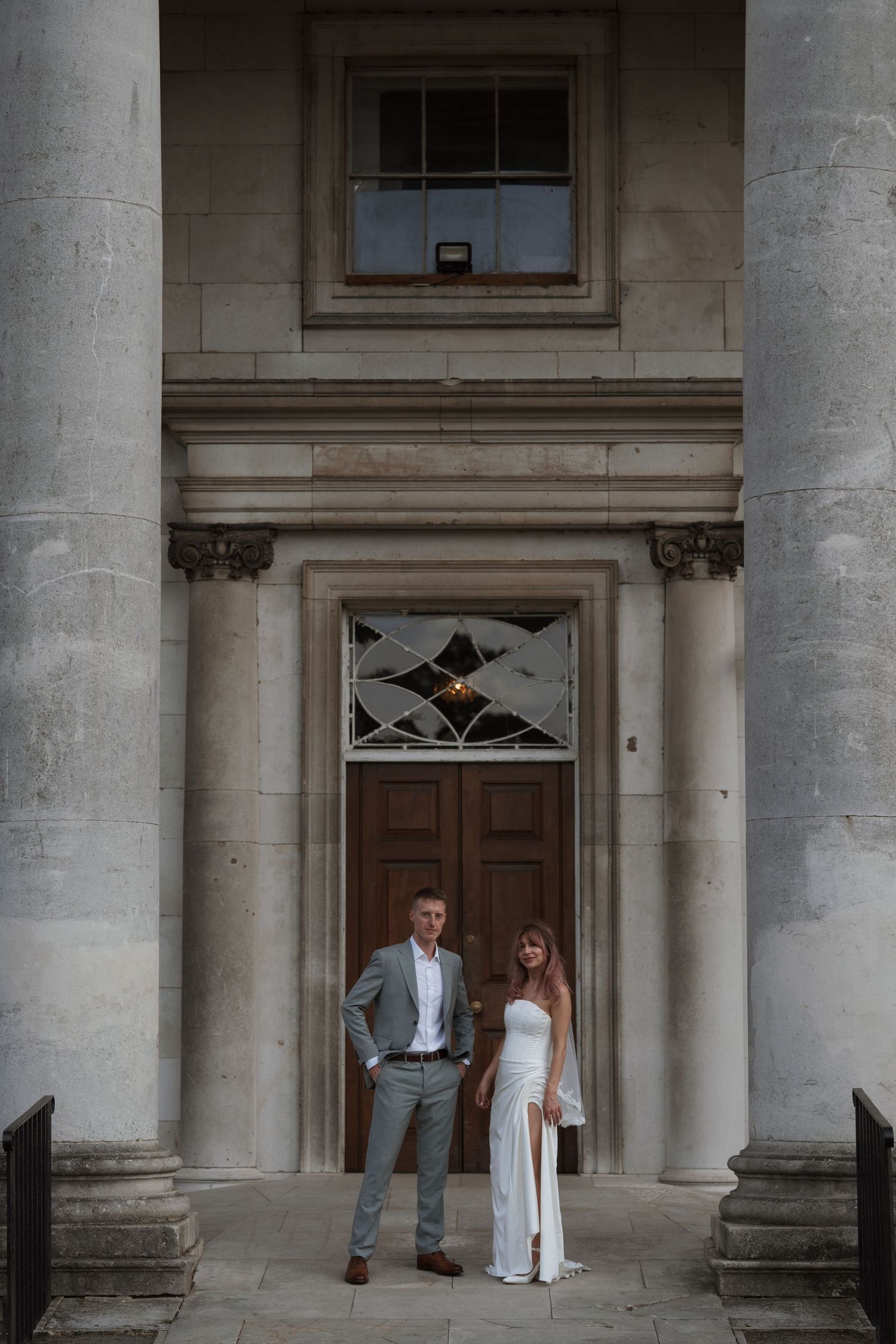 A couple in wedding attire stands between elegant stone columns of a historic building.