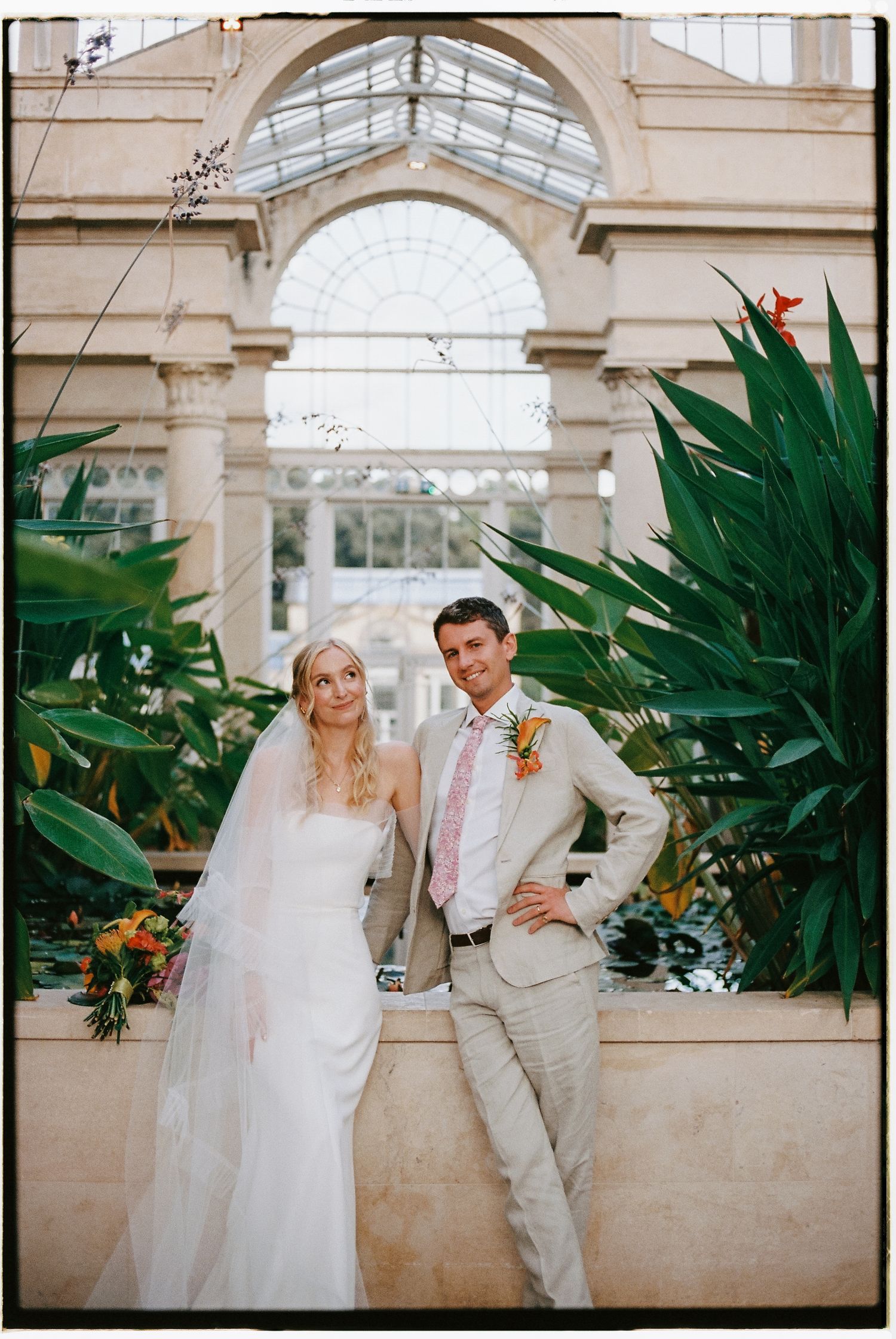 Wedding couple poses in front of a conservatory with tropical plants and archways.