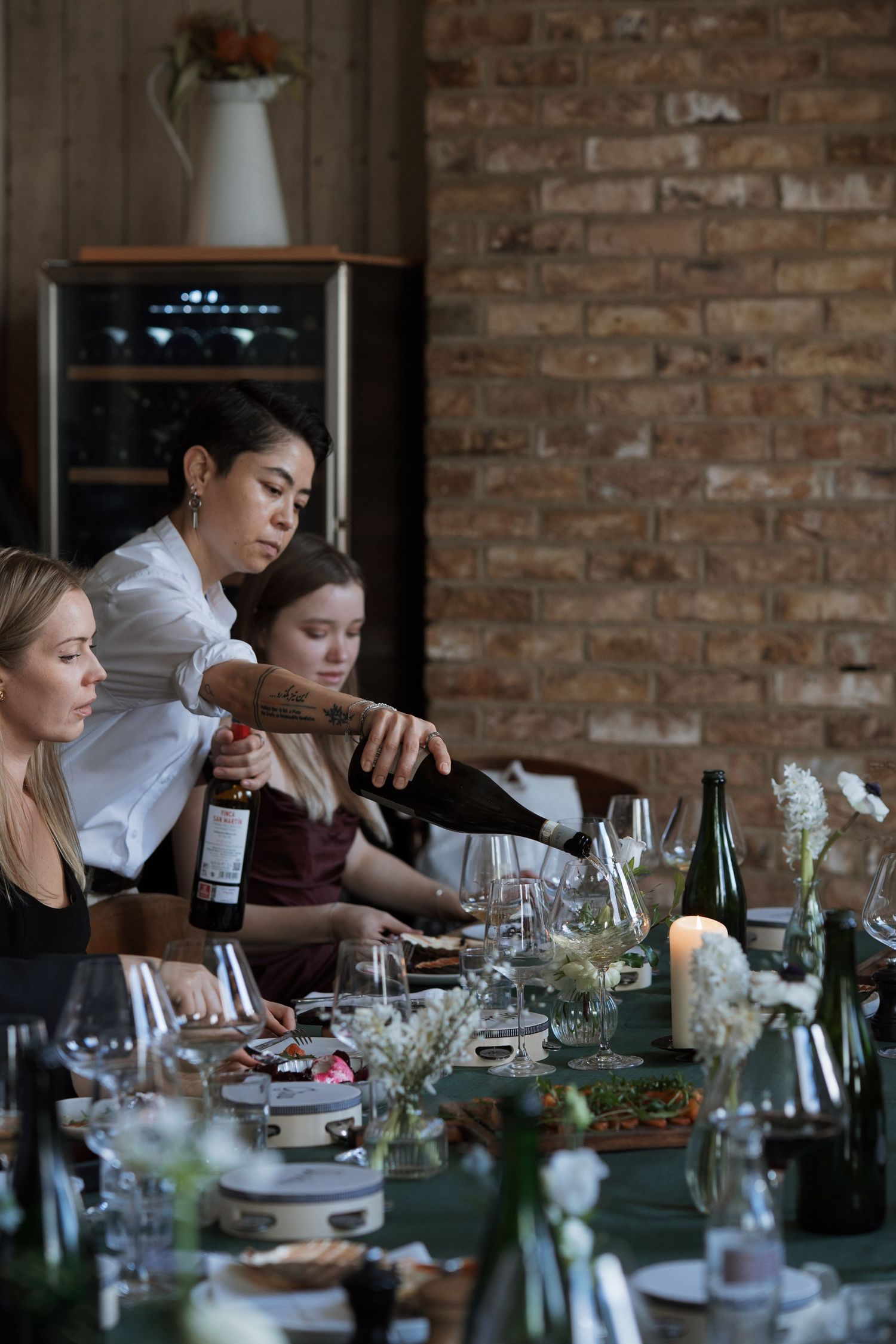 Diners gathered around a table with wine bottles and floral centerpieces in a rustic brick setting.