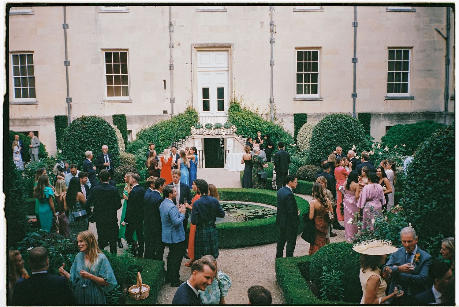 Elegant outdoor wedding reception in a formal garden courtyard with guests mingling among sculpted hedges.