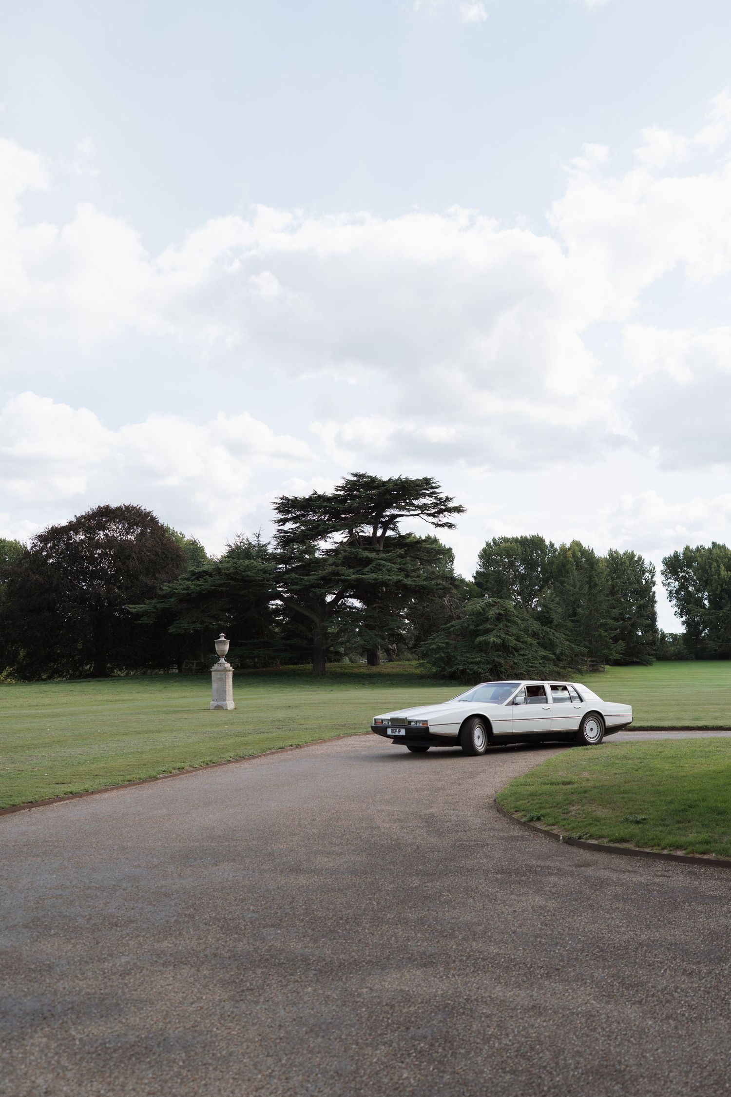A vintage white car parked on a gravel driveway overlooking expansive green grounds.