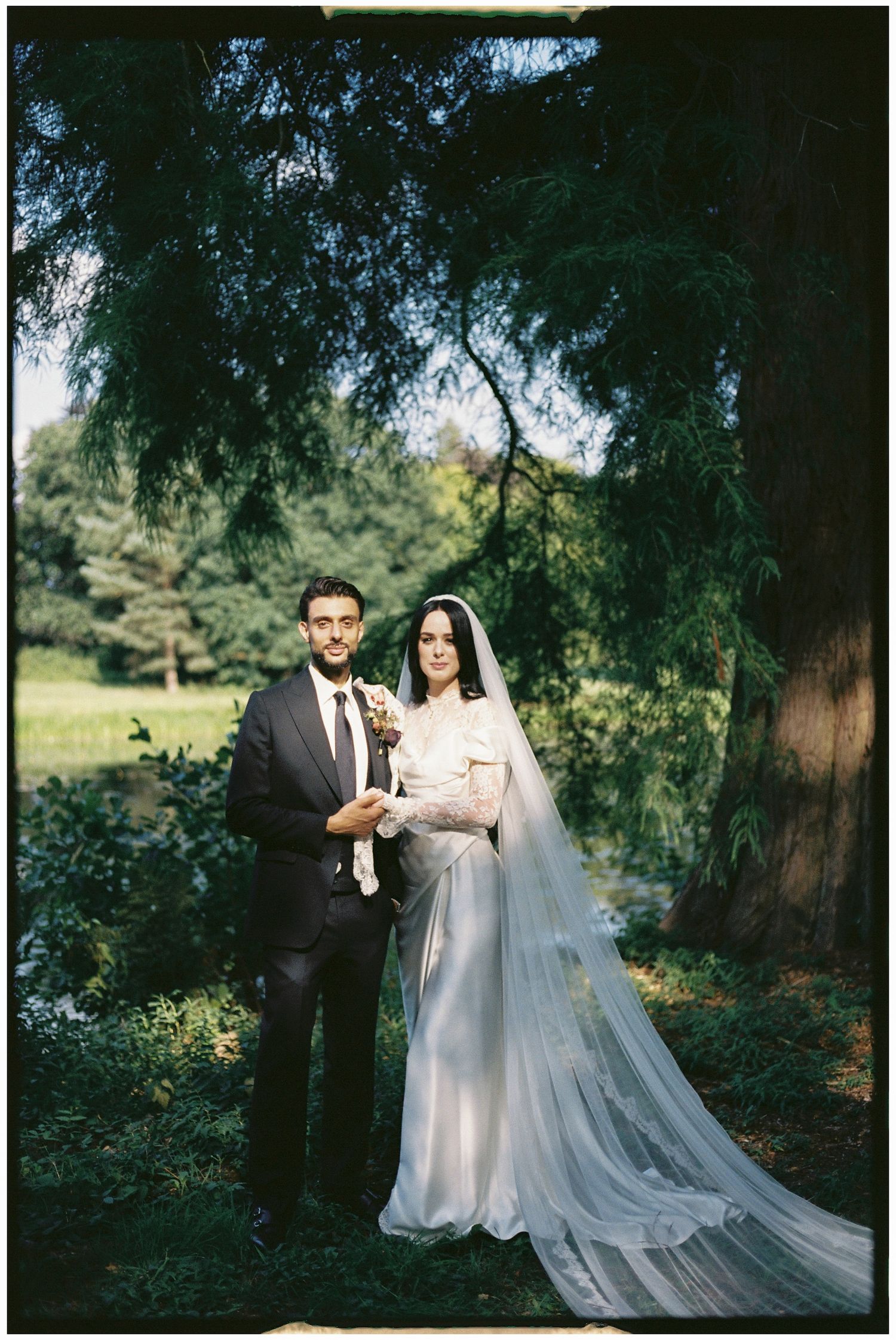 Newlyweds sharing intimate moment under trees with flowing veil.