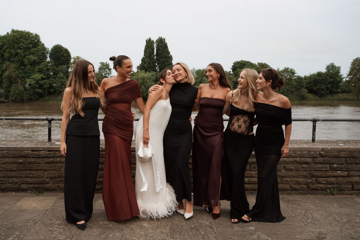 Wedding party in burgundy and black dresses poses along stone wall outdoors.