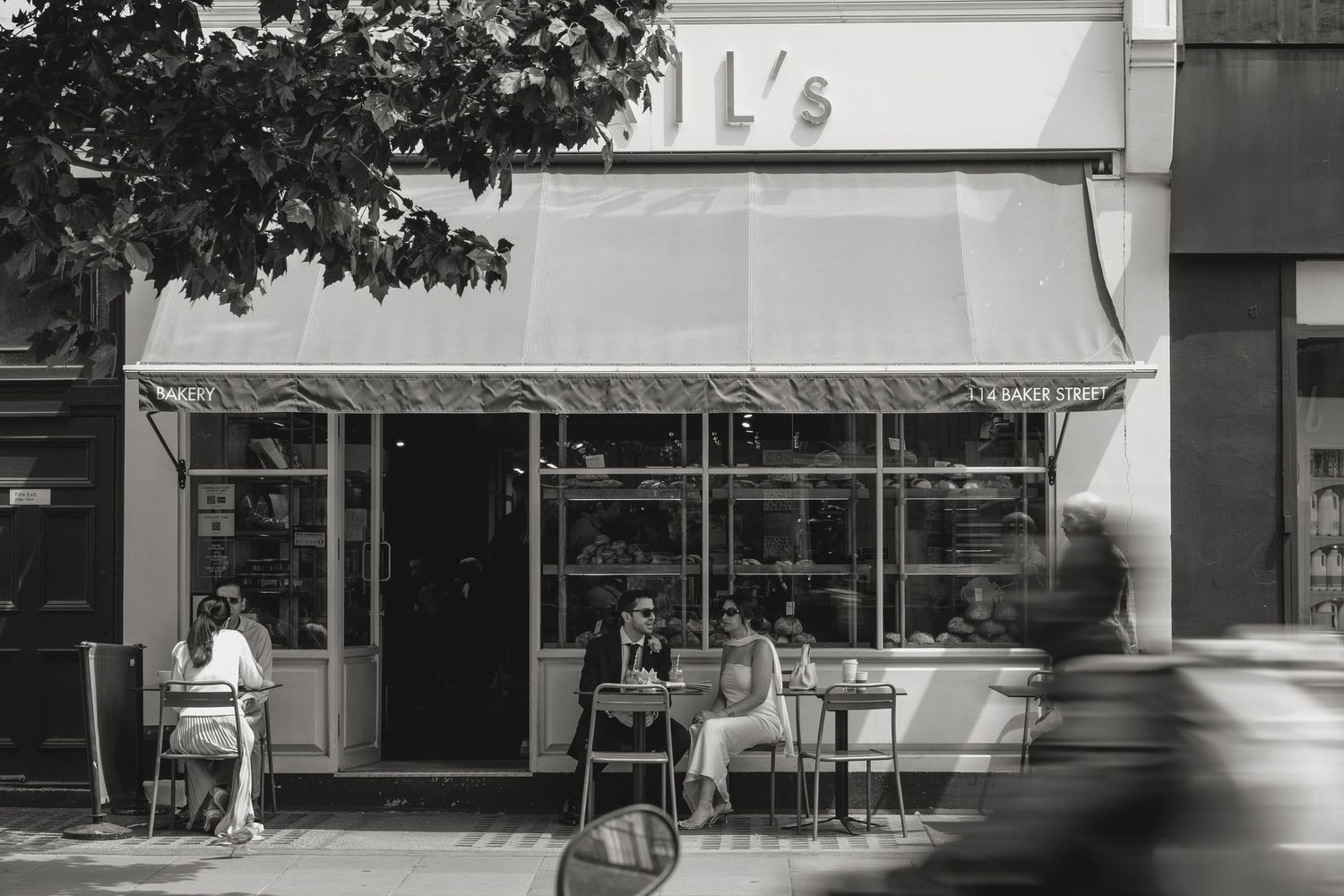 Black and white photo of a cozy street cafe with outdoor seating, patrons dining under an awning on a sunny day.