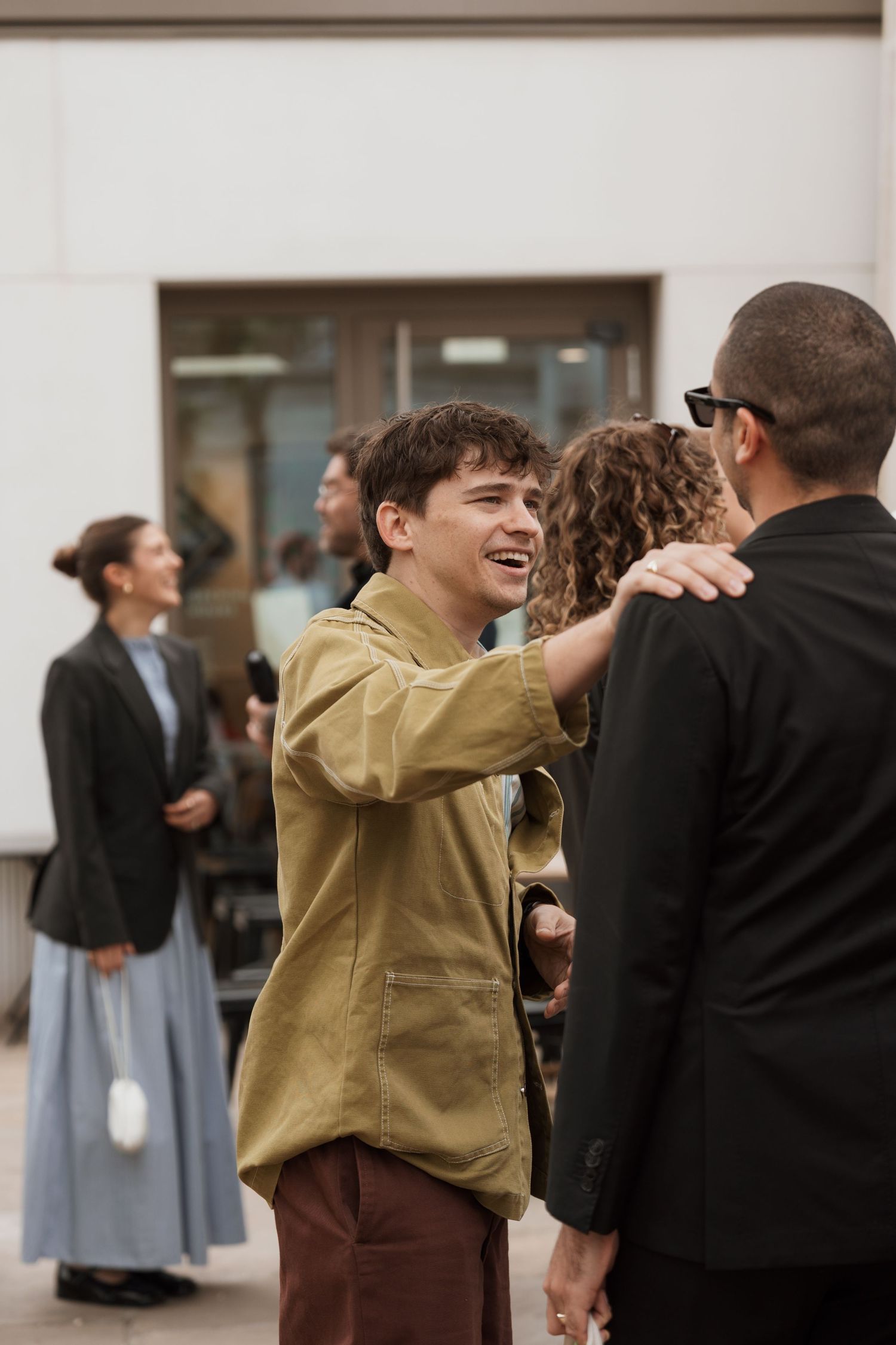Young man in olive shirt smiling and greeting friends at an outdoor social gathering.