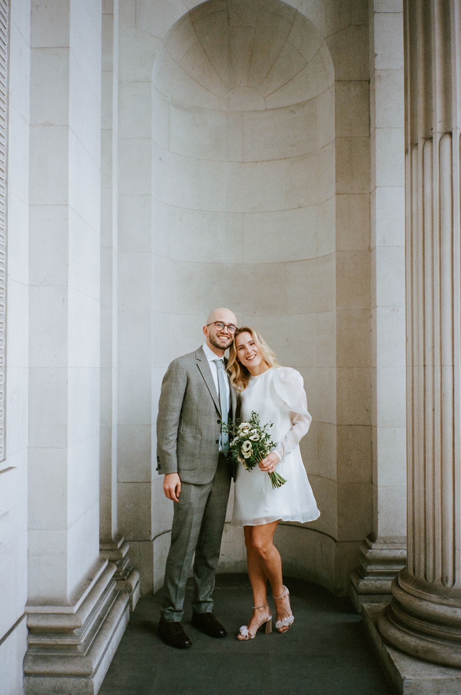 Bride in white mini dress and groom in gray suit pose between stone columns holding a greenery bouquet.