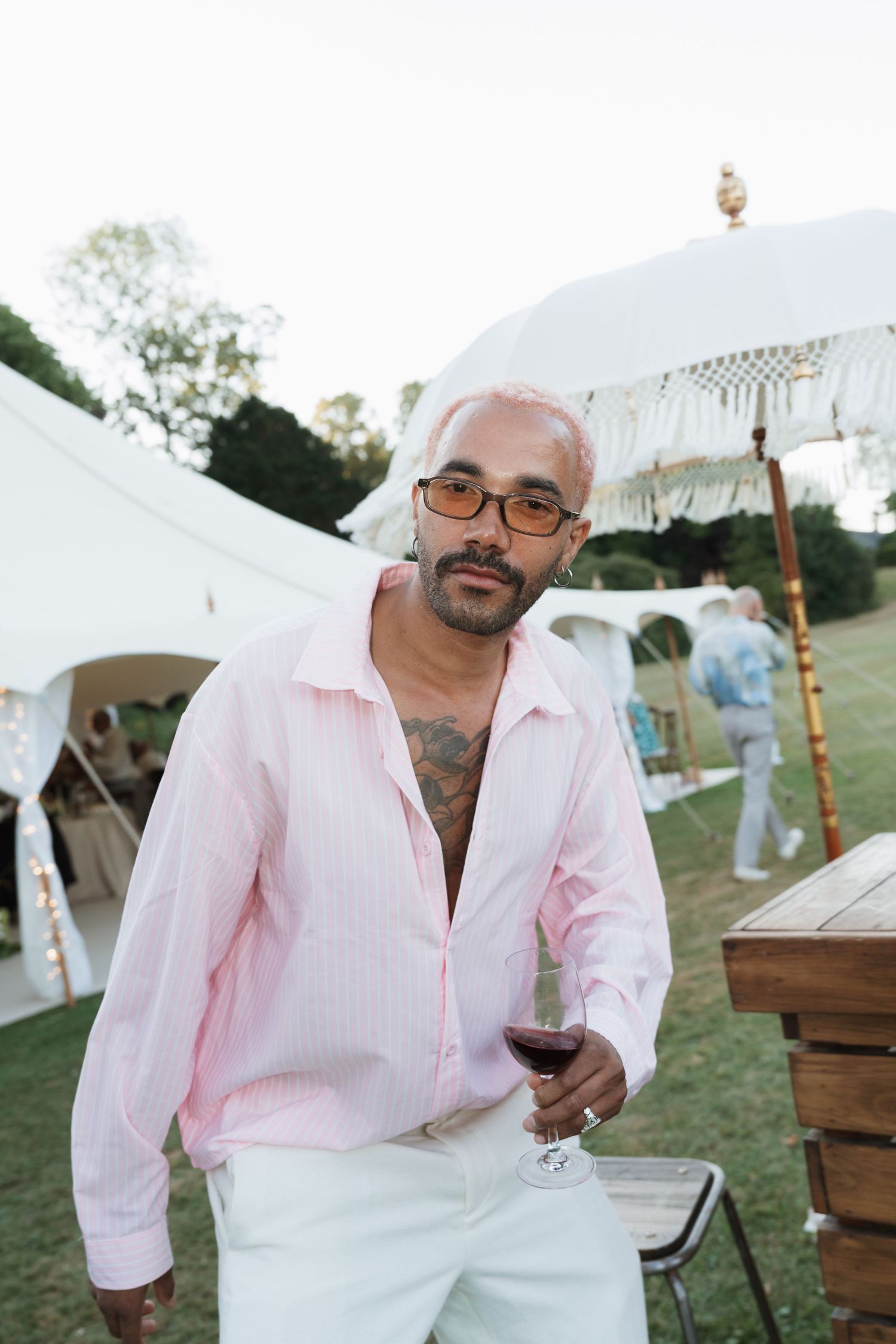 Man in pink shirt holding red wine glass at outdoor garden party with white tent.