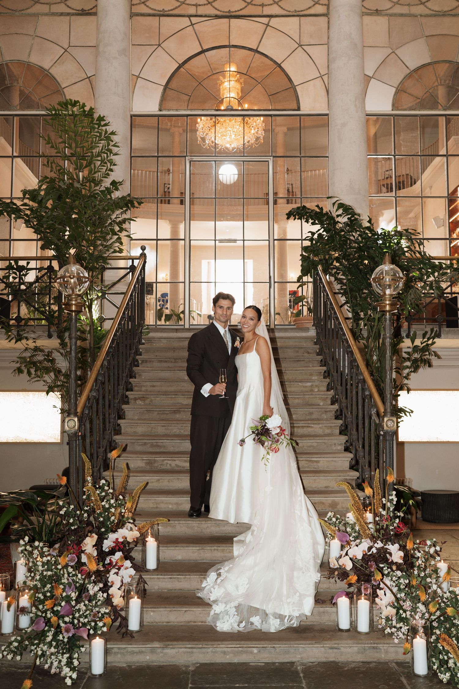 Bride in a flowing white gown and groom in a black suit pose on grand stone stairs surrounded by candles, flowers and tall windows.