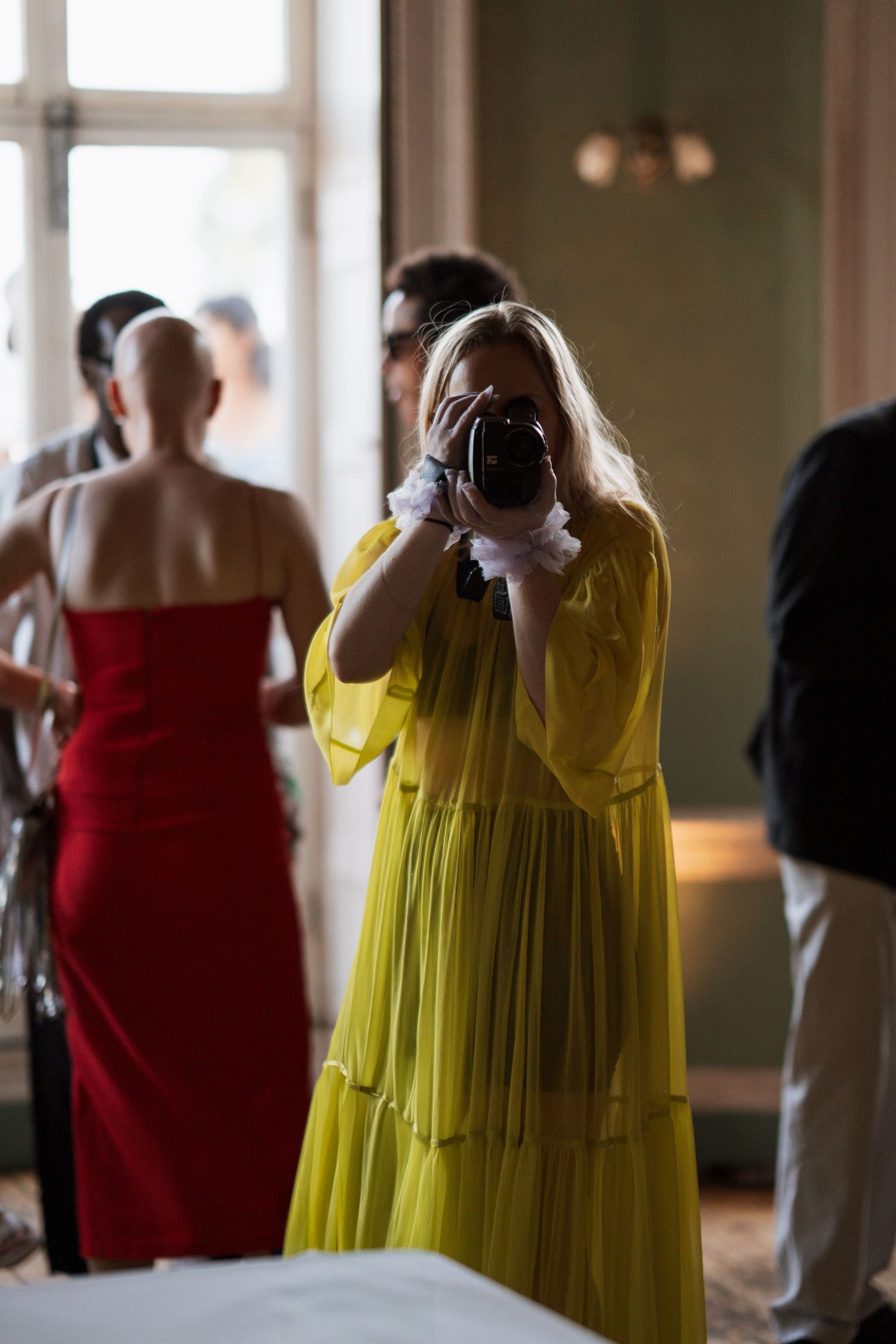 Wedding attendees in bright yellow and red dresses taking photos by large windows.