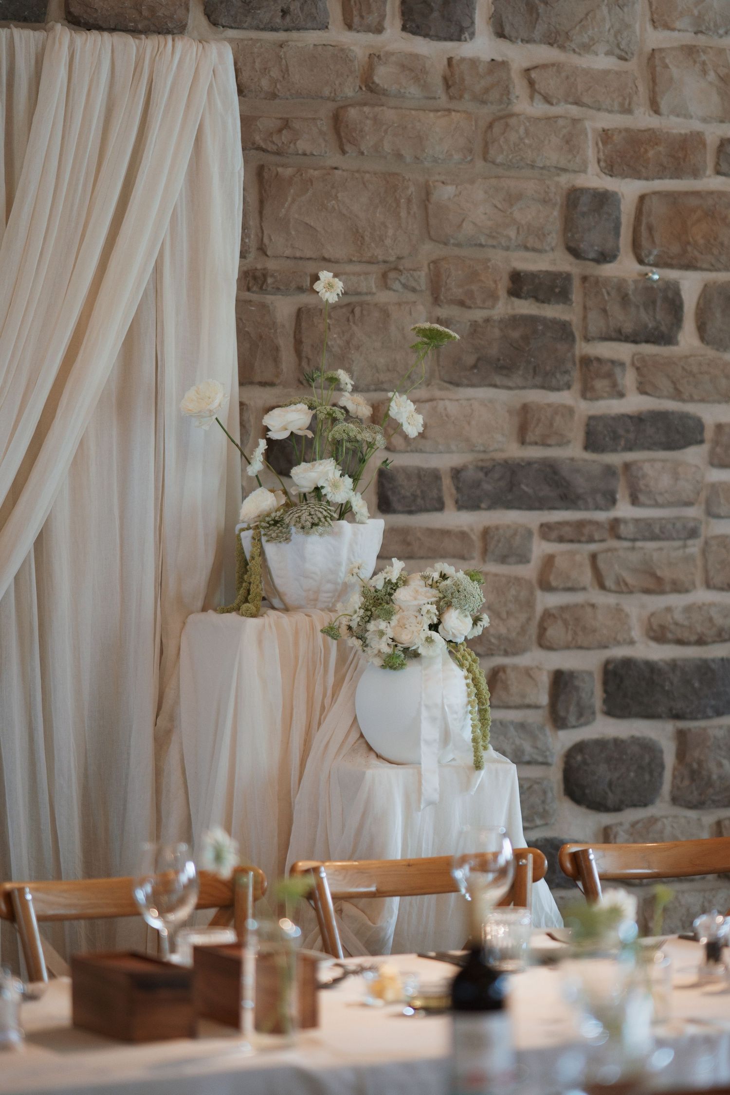 Wedding reception decor featuring white floral arrangements against rustic stone wall.