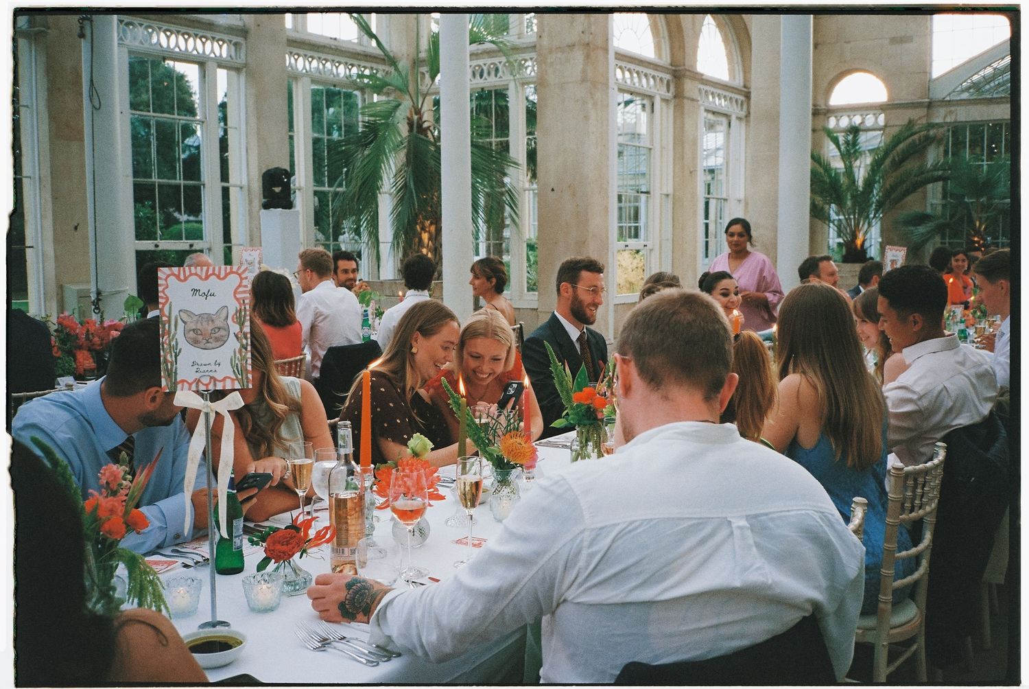Wedding reception guests seated at elegant tables in a bright conservatory setting.