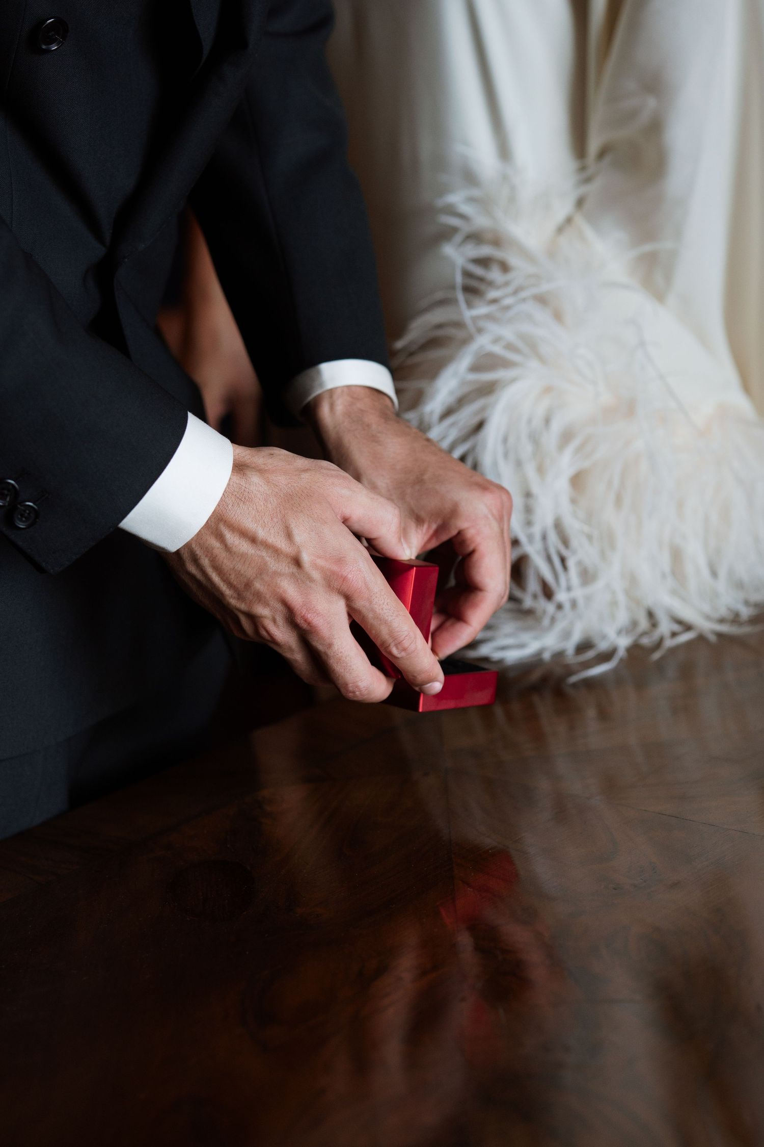 Close-up of hands near a white feathered dress and black suit jacket on a polished wooden surface.