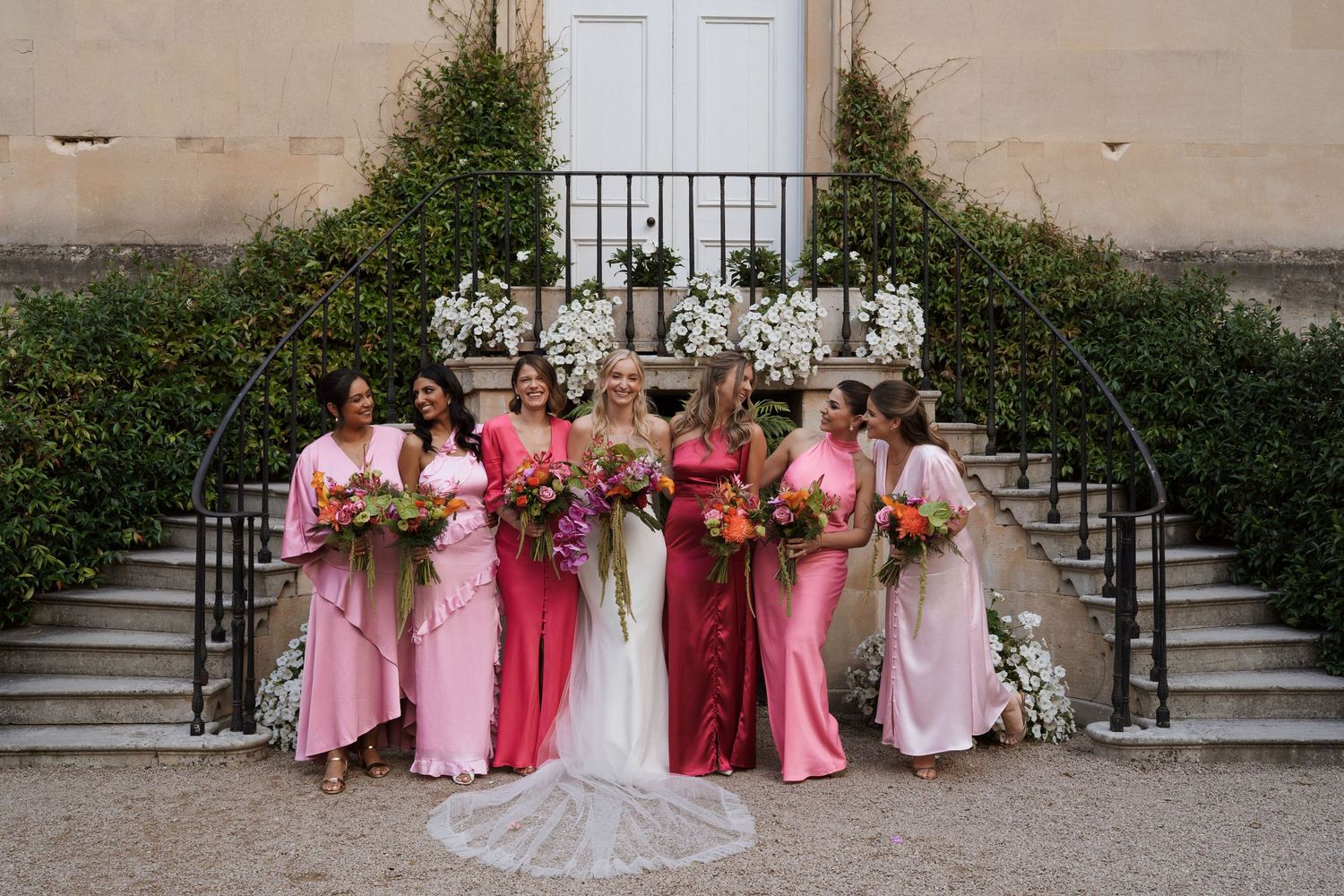 Bridesmaids in pink dresses pose with the bride on garden steps.