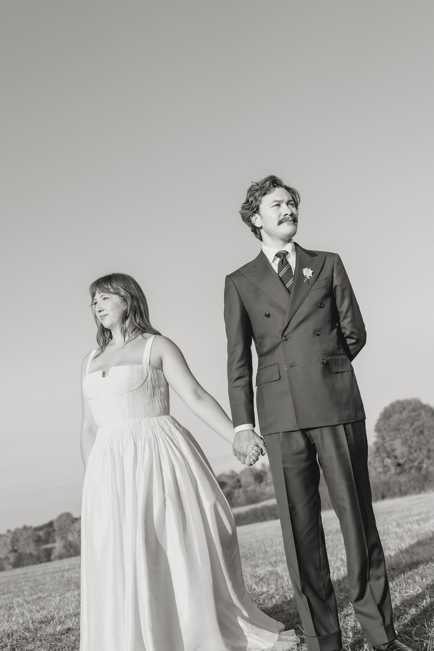 Black and white photo of a bride and groom holding hands outdoors in a field on their wedding day.