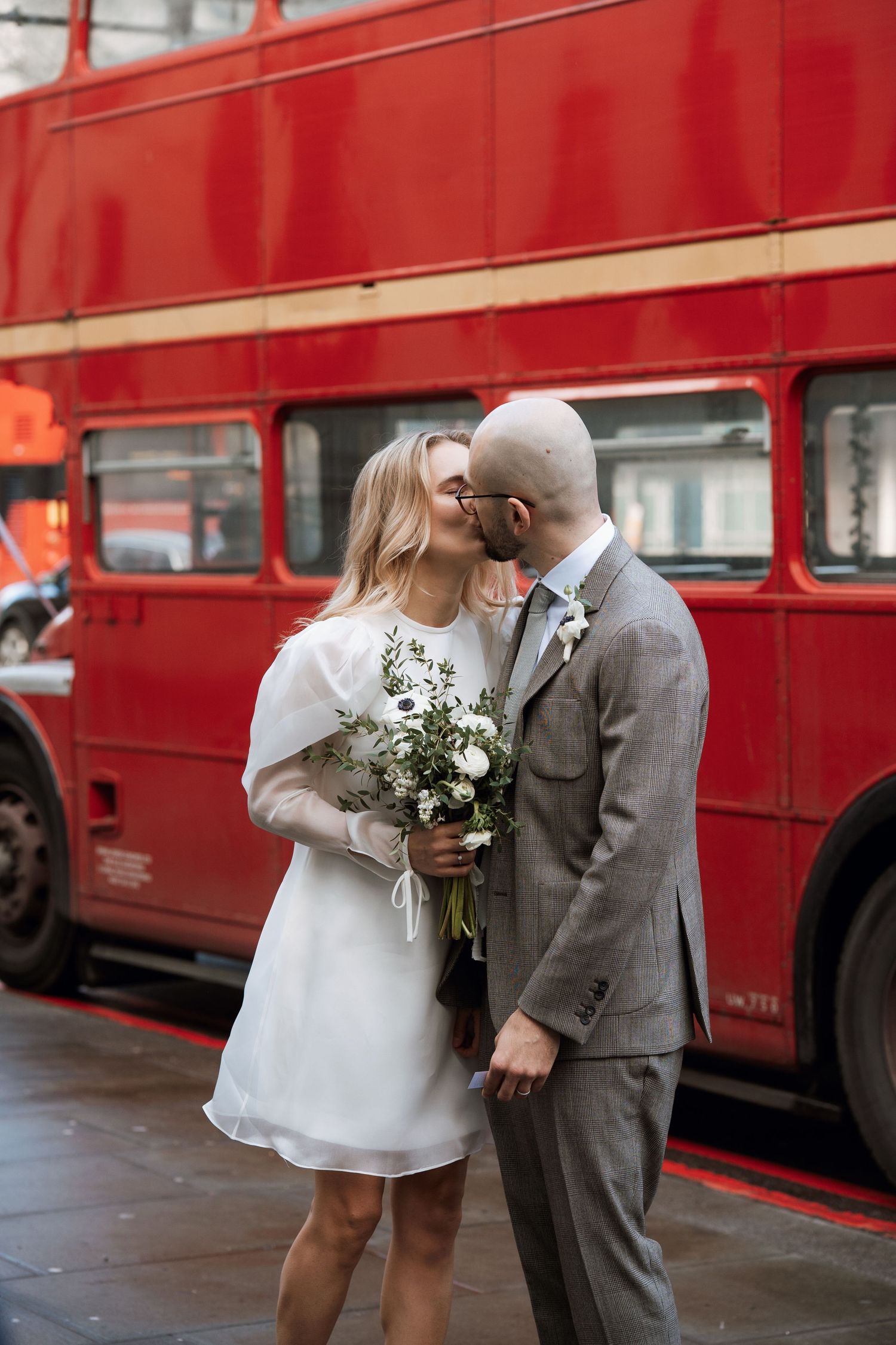 A couple shares an intimate moment in front of a red London bus, with the bride holding a white floral bouquet.