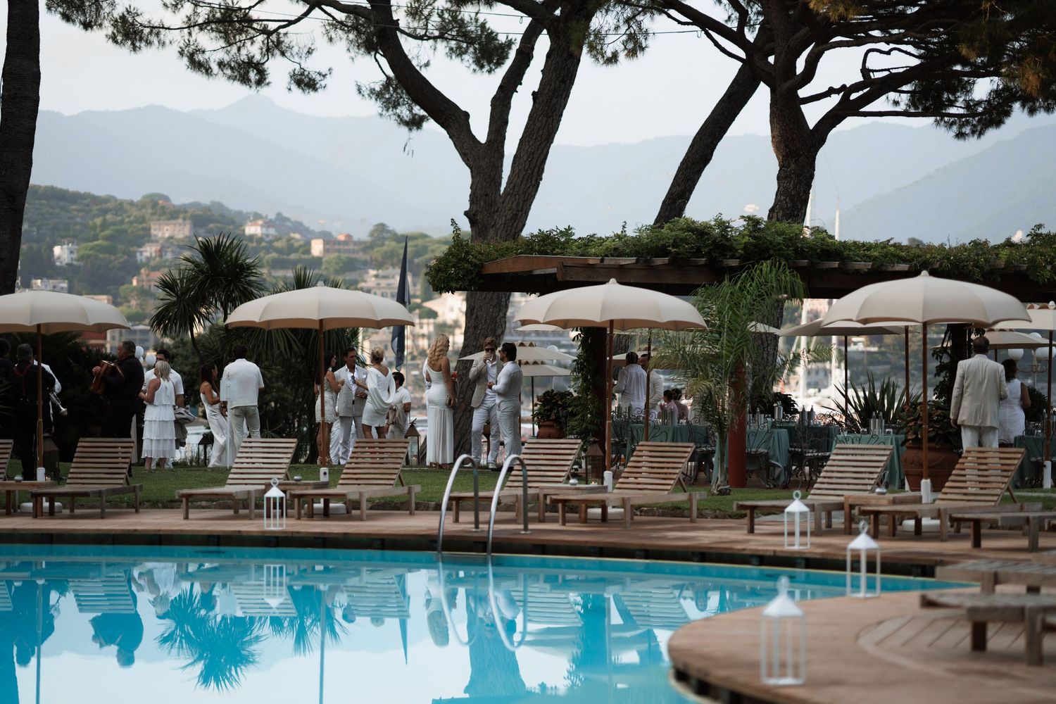 Elegant poolside setting at a luxury hotel with white umbrellas, lounge chairs, and guests in white attire.