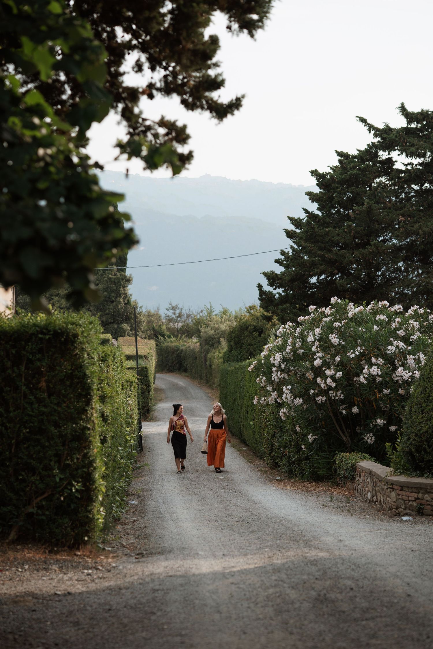Two women walking down a scenic gravel path lined with hedges and flowering trees in a Mediterranean landscape.
