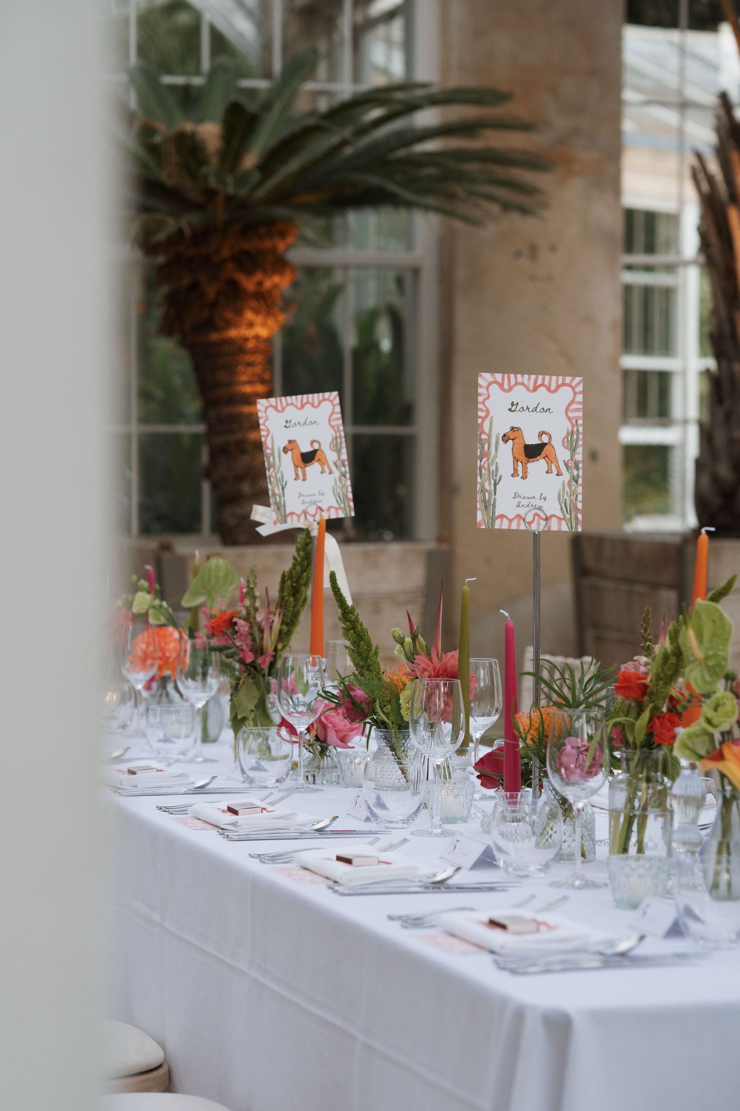 Long white reception table decorated with floral arrangements and place settings under palm trees.