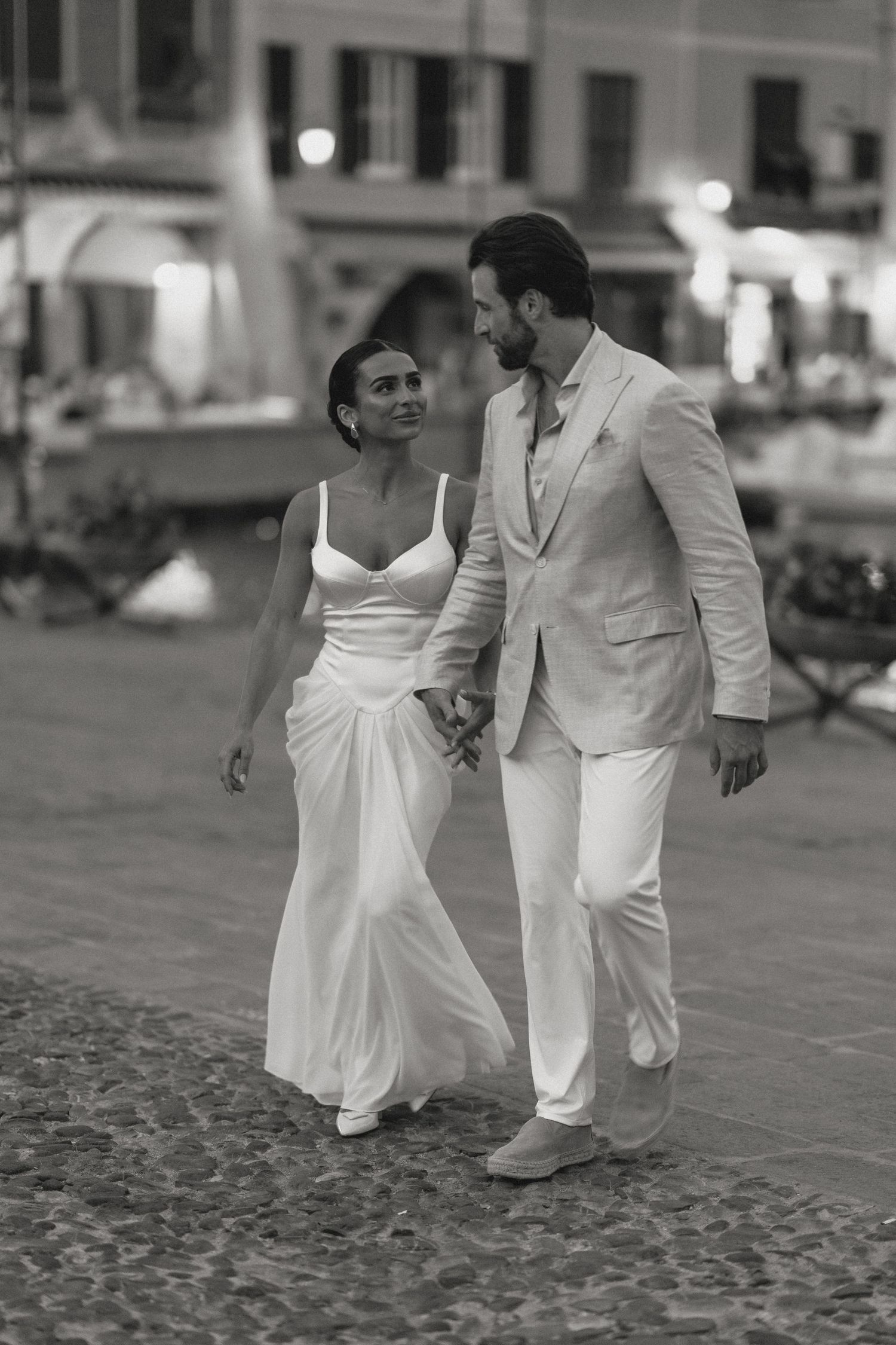 Black and white photo of a couple walking on cobblestone streets in formal attire.