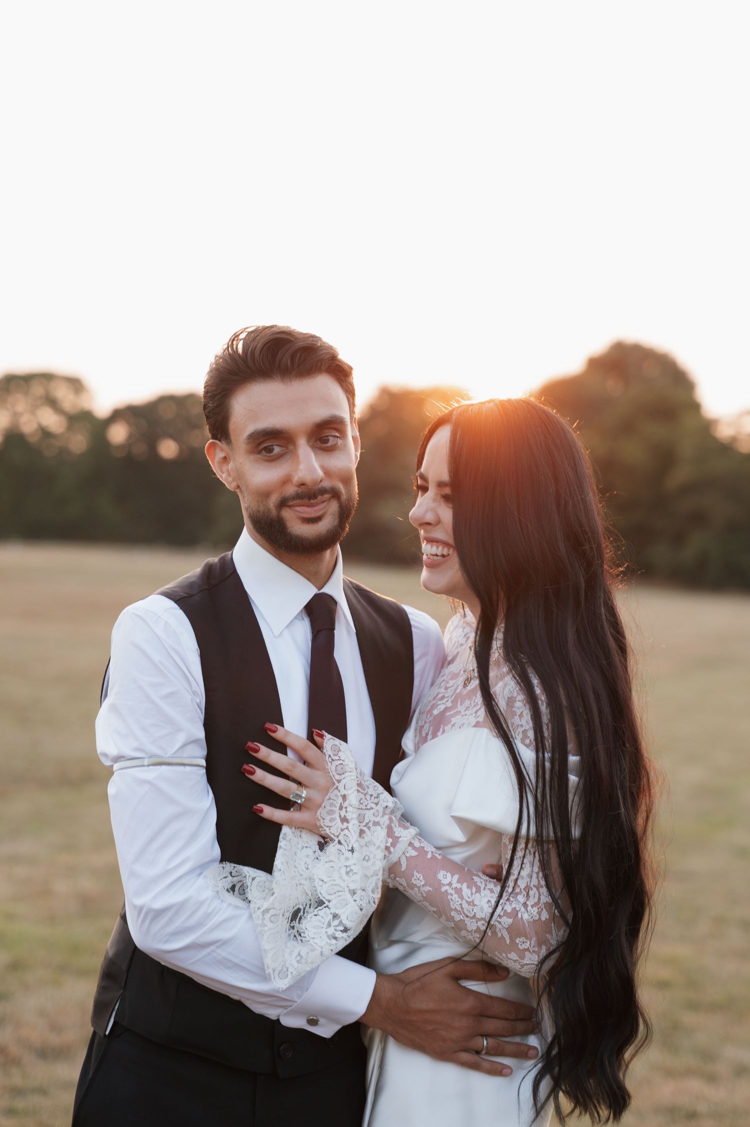 A romantic couple embraces in a field during golden hour wearing formal wedding attire.