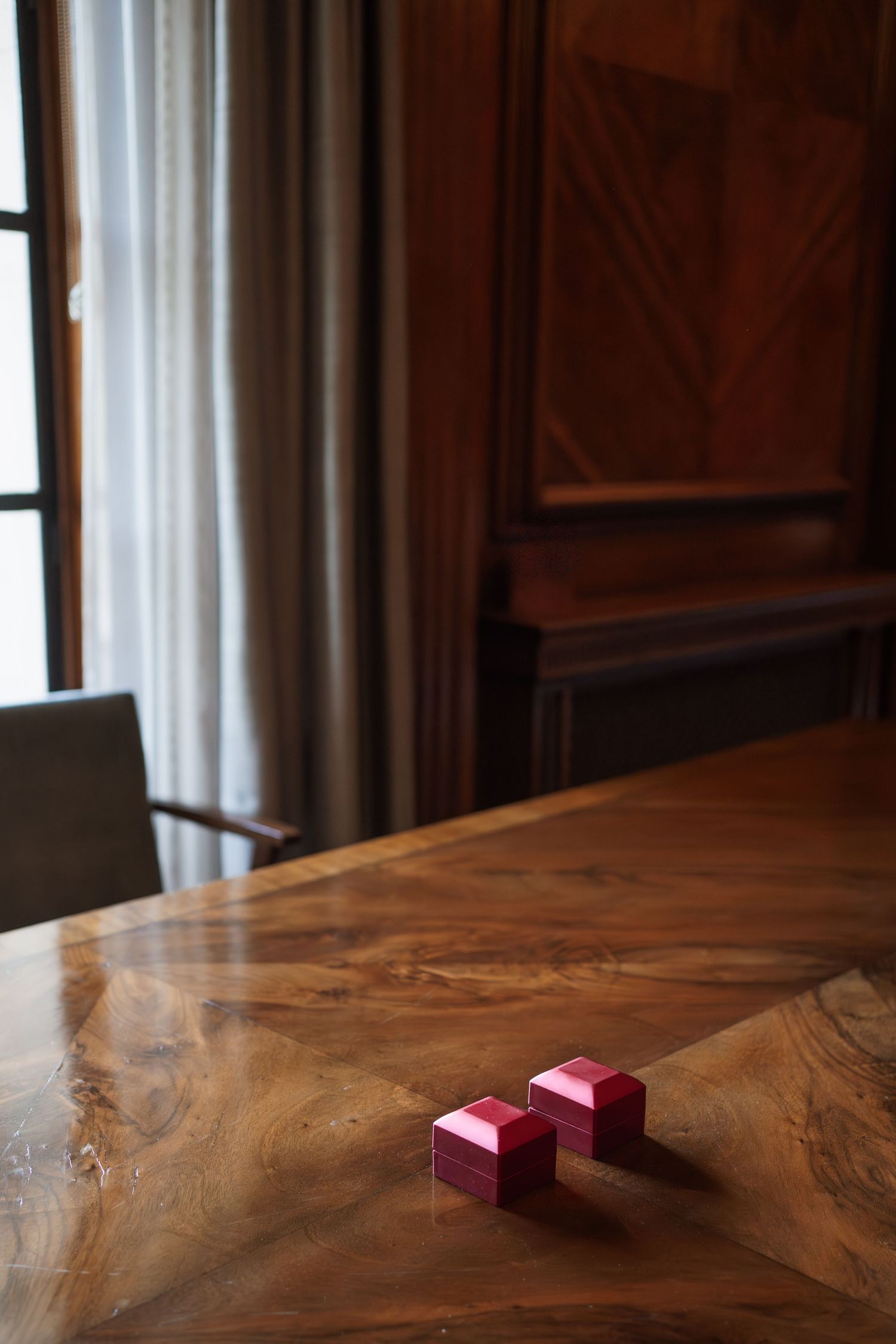 Two red ring boxes on a polished wooden table near a window with elegant curtains.