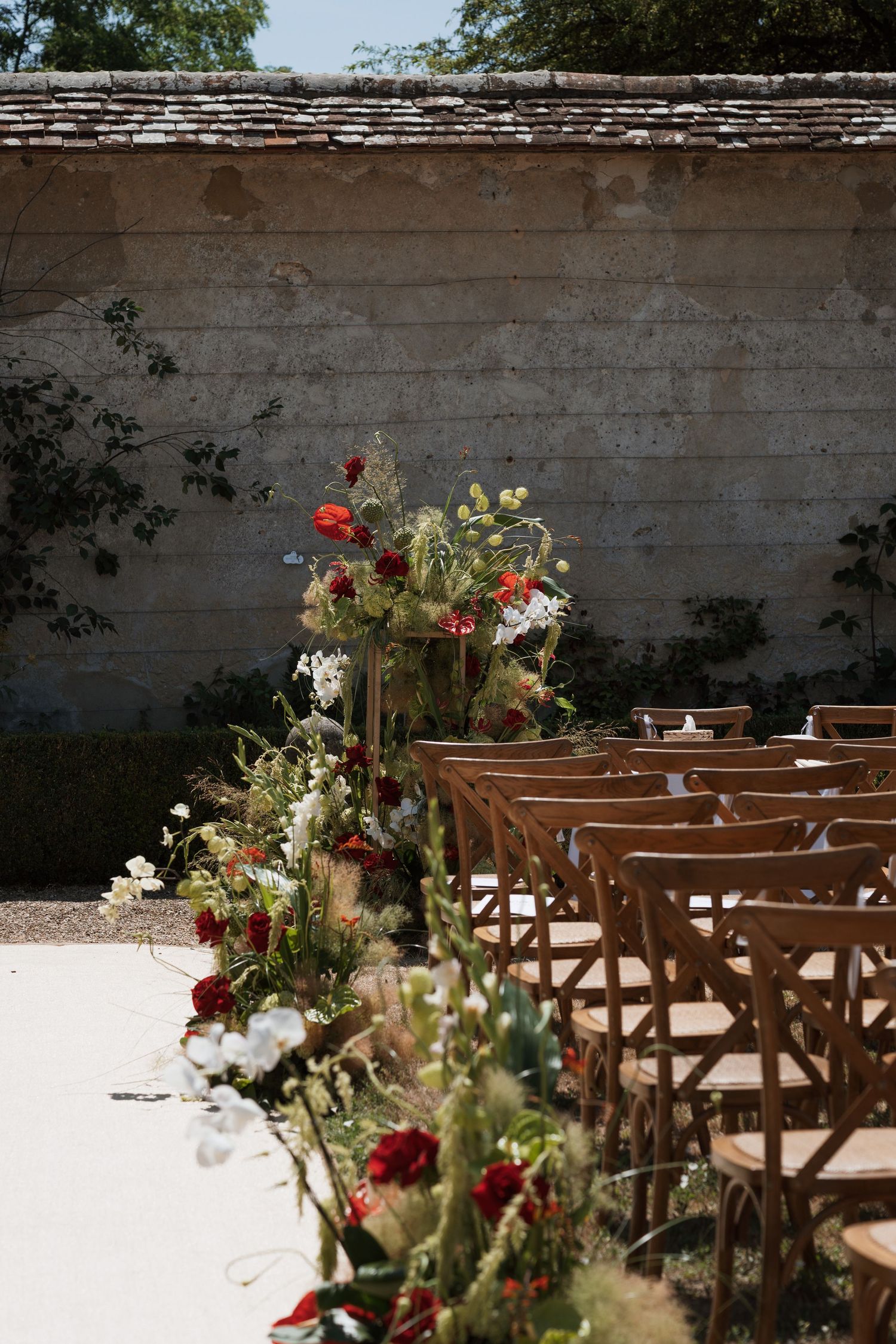 Outdoor wedding ceremony setup with wooden cross-back chairs, red and white floral aisle arrangements, and a rustic stone wall backdrop.